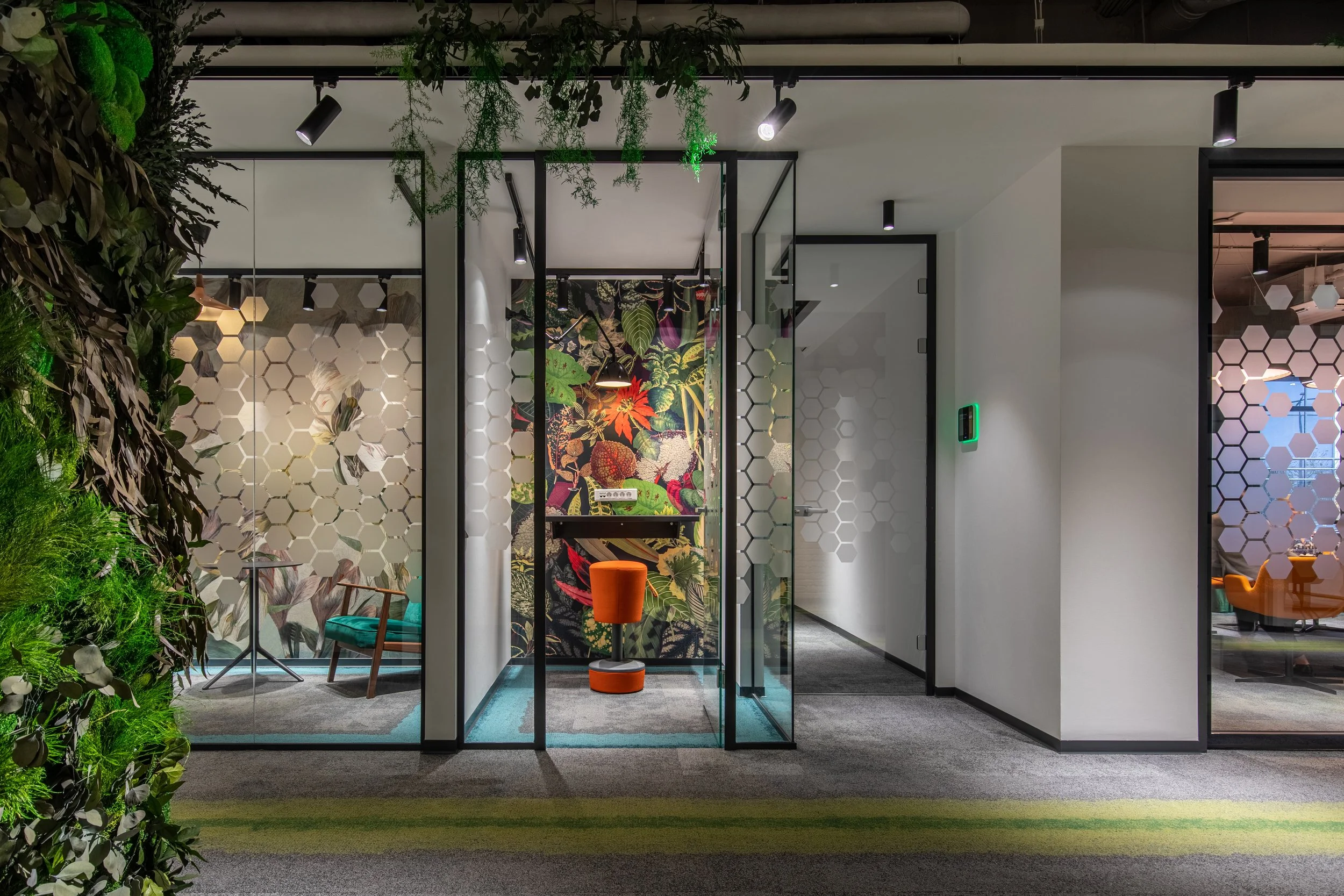 Modern office space with glass-walled conference rooms decorated with vibrant wallpaper and greenery, featuring chairs, a small table, and a workspace with a green wall panel.