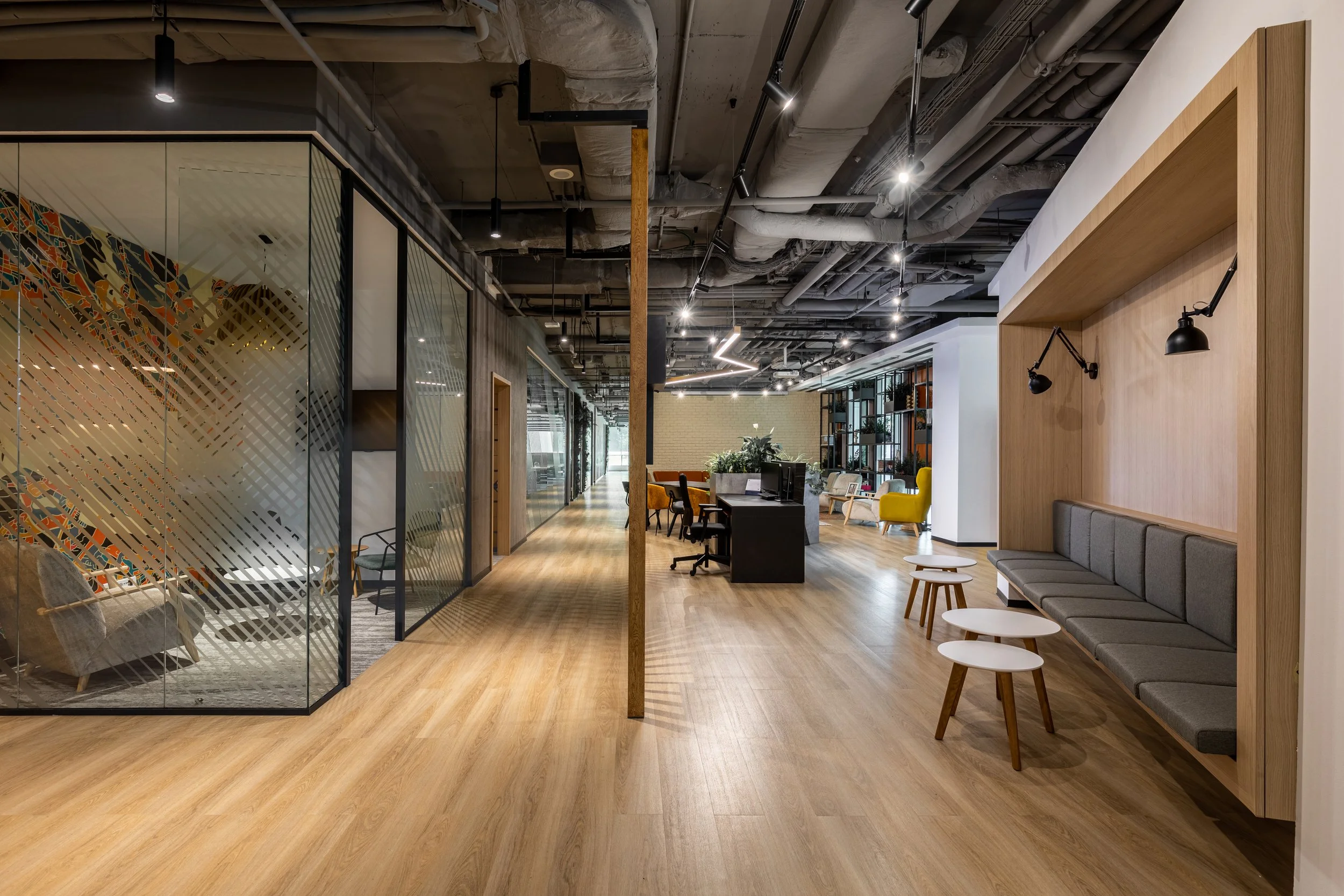Modern office lounge area with wooden flooring, a large sectional gray sofa with white and wooden tables, and a wall with black adjustable reading lamps. There are glass-walled rooms on the left and a work area with chairs and desks in the background. The ceiling has exposed ducts and pipes with track lighting.