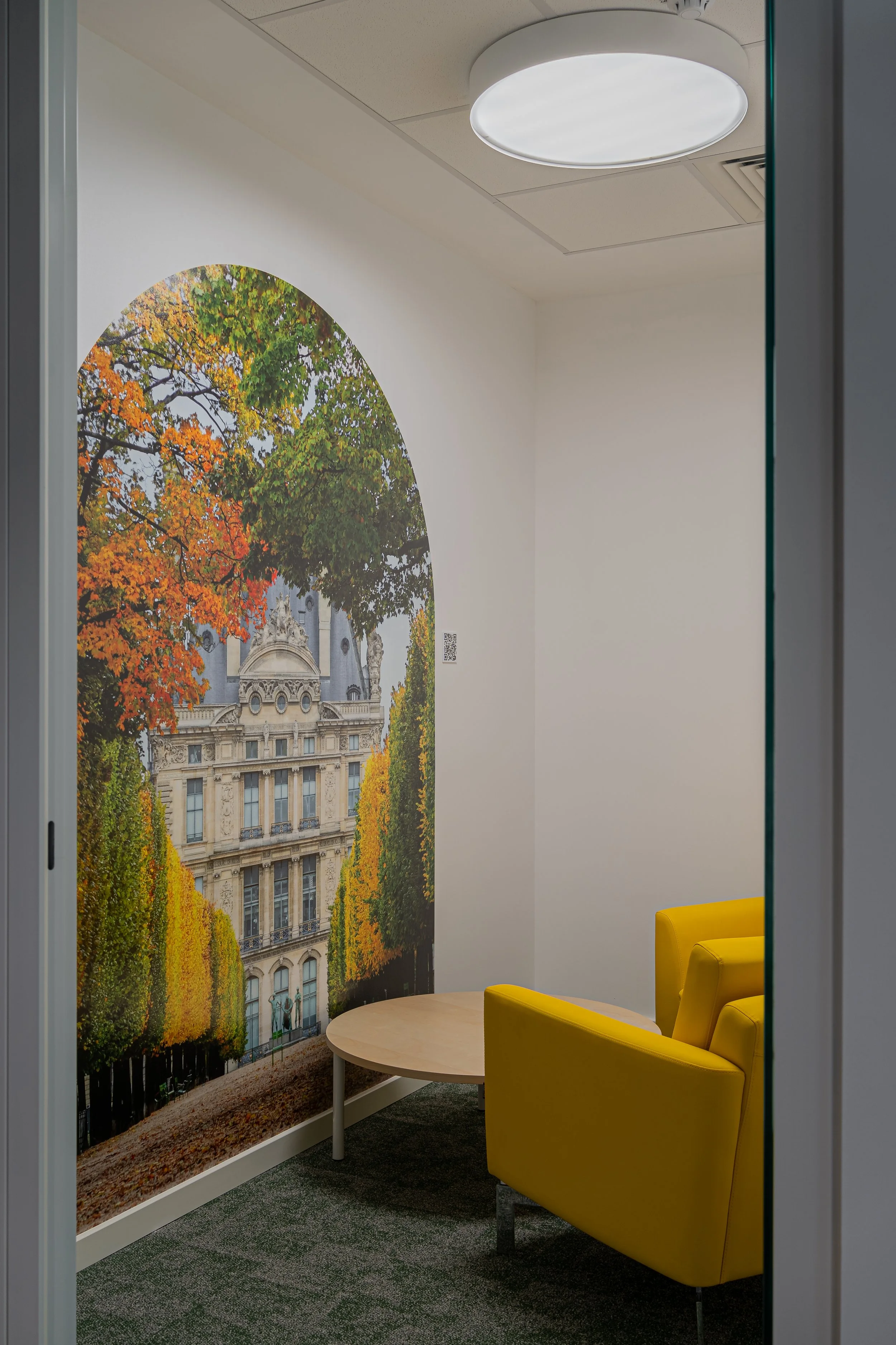 A small room with white walls, a yellow armchair, a round wooden table, and a wall mural of a historic building surrounded by fall foliage.