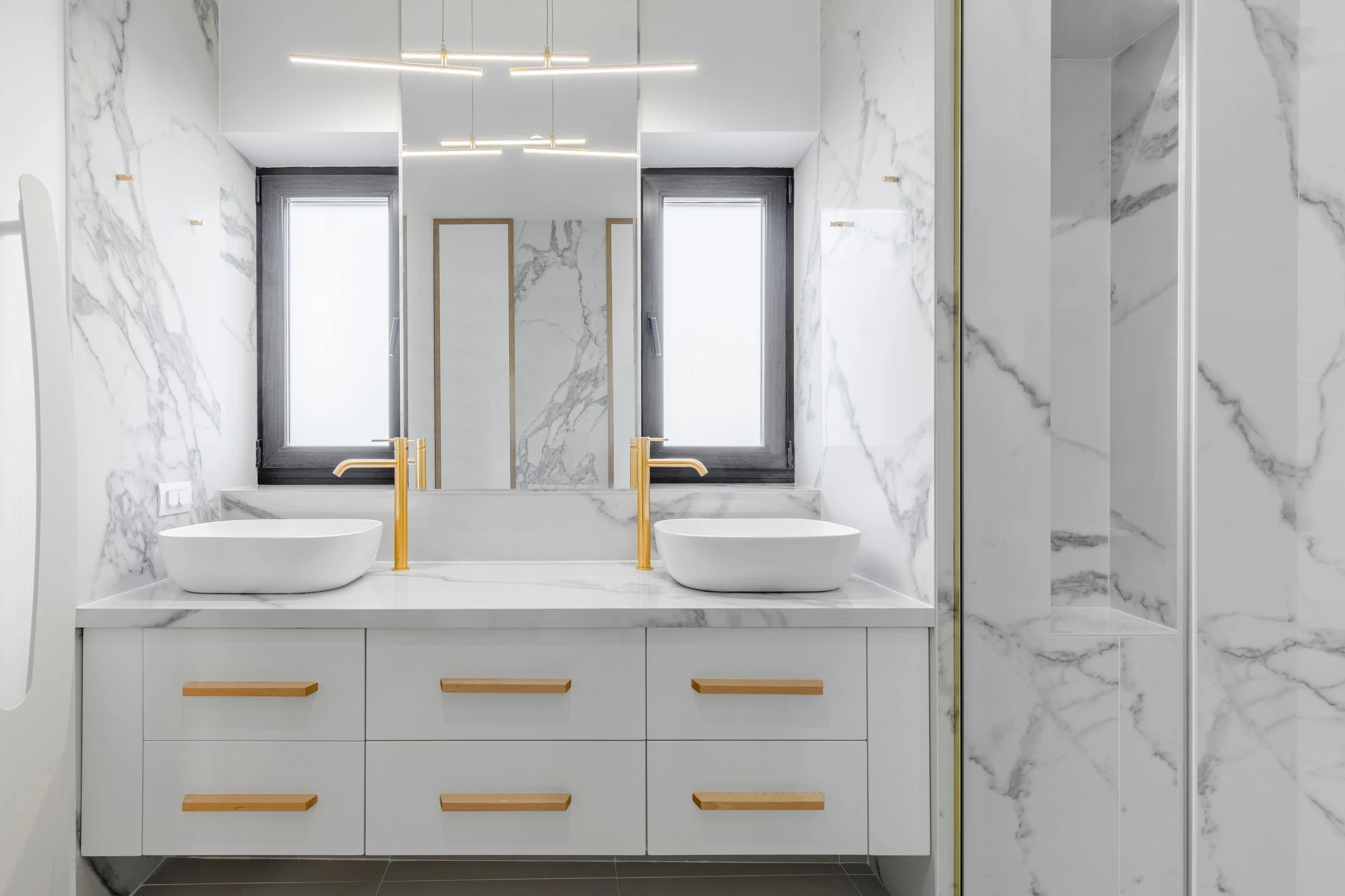 Modern bathroom with white marble walls, two square vessel sinks with gold faucets, a large mirror, black-framed windows, and a shower enclosure.