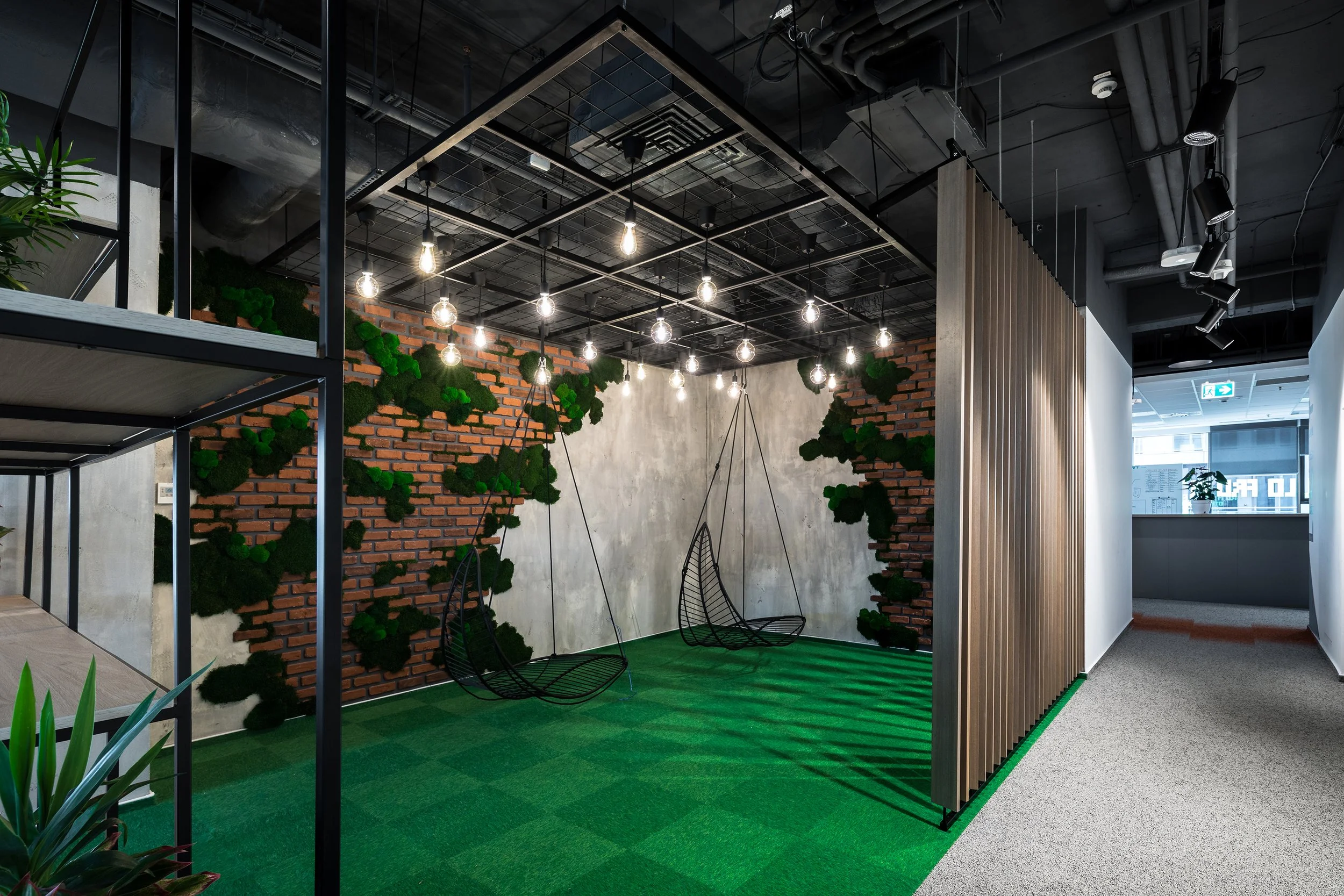 Modern office lounge with hanging light bulbs, green moss wall decor, black metal swings, and a partial wooden partition.