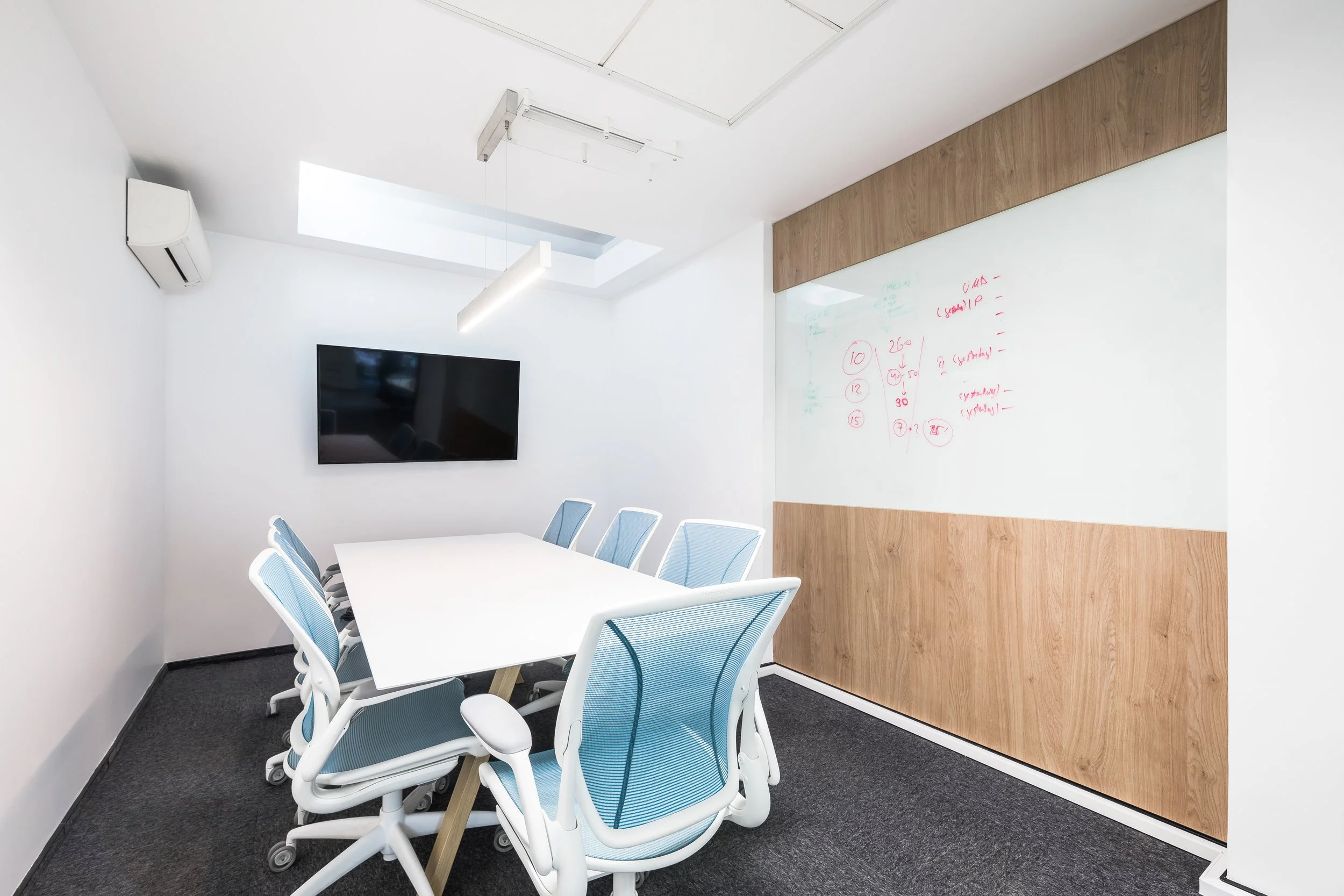 Empty modern conference room with a white table, blue and white office chairs, a wall-mounted flat-screen TV, a whiteboard with red markings, a wood accent wall, and a ceiling with a skylight and hanging light fixture.