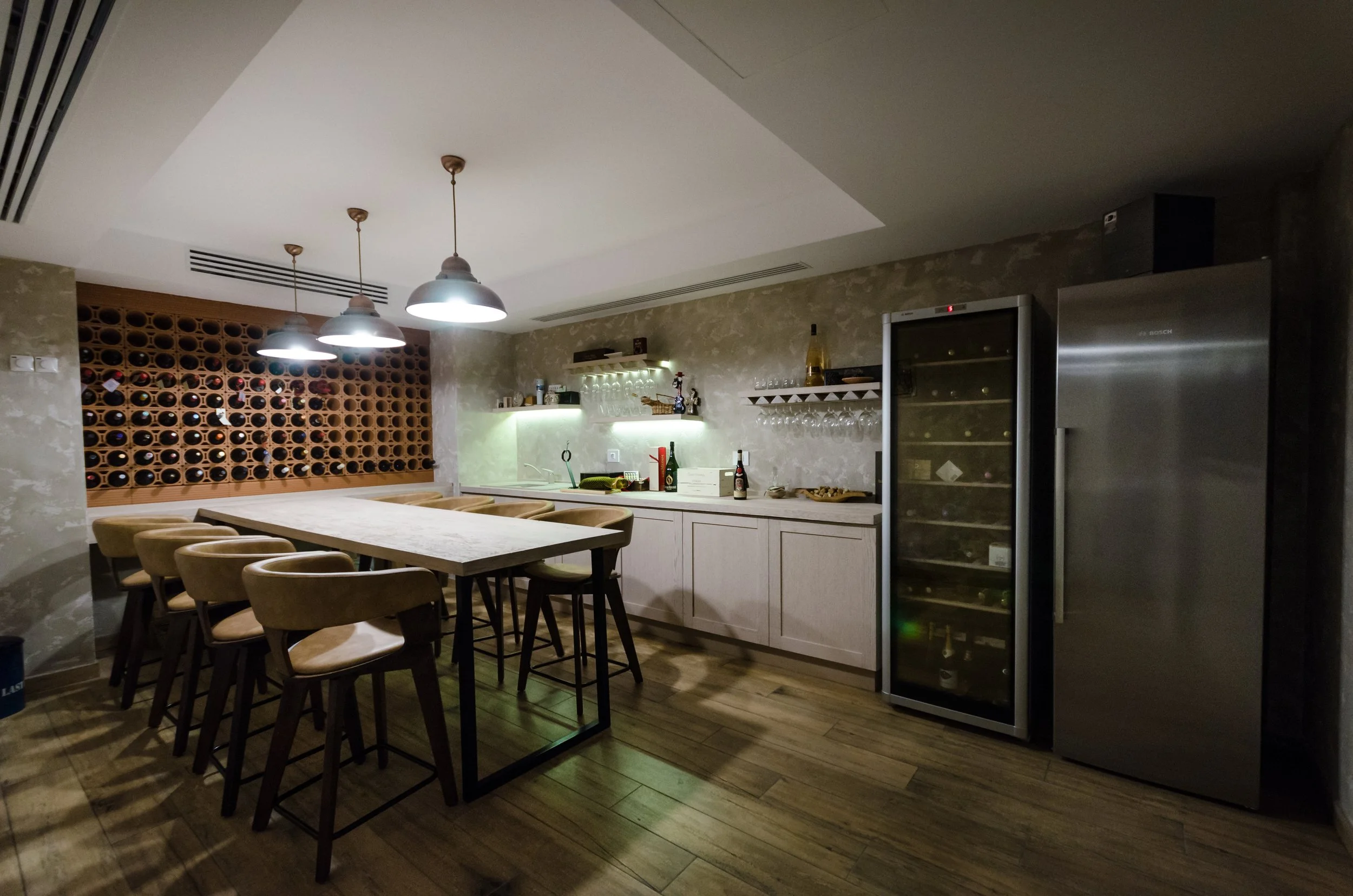 Modern wine bar or kitchen with a long table and beige chairs, wine bottles on a wall rack, shelving with wine glasses, a wine fridge, and a stainless steel refrigerator.