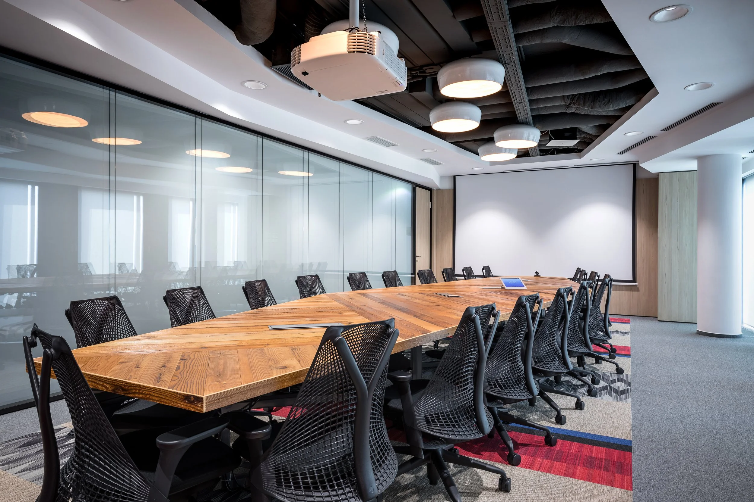 A modern conference room with a large wooden table, black mesh chairs, a projector ceiling mount, and a large white screen on the back wall