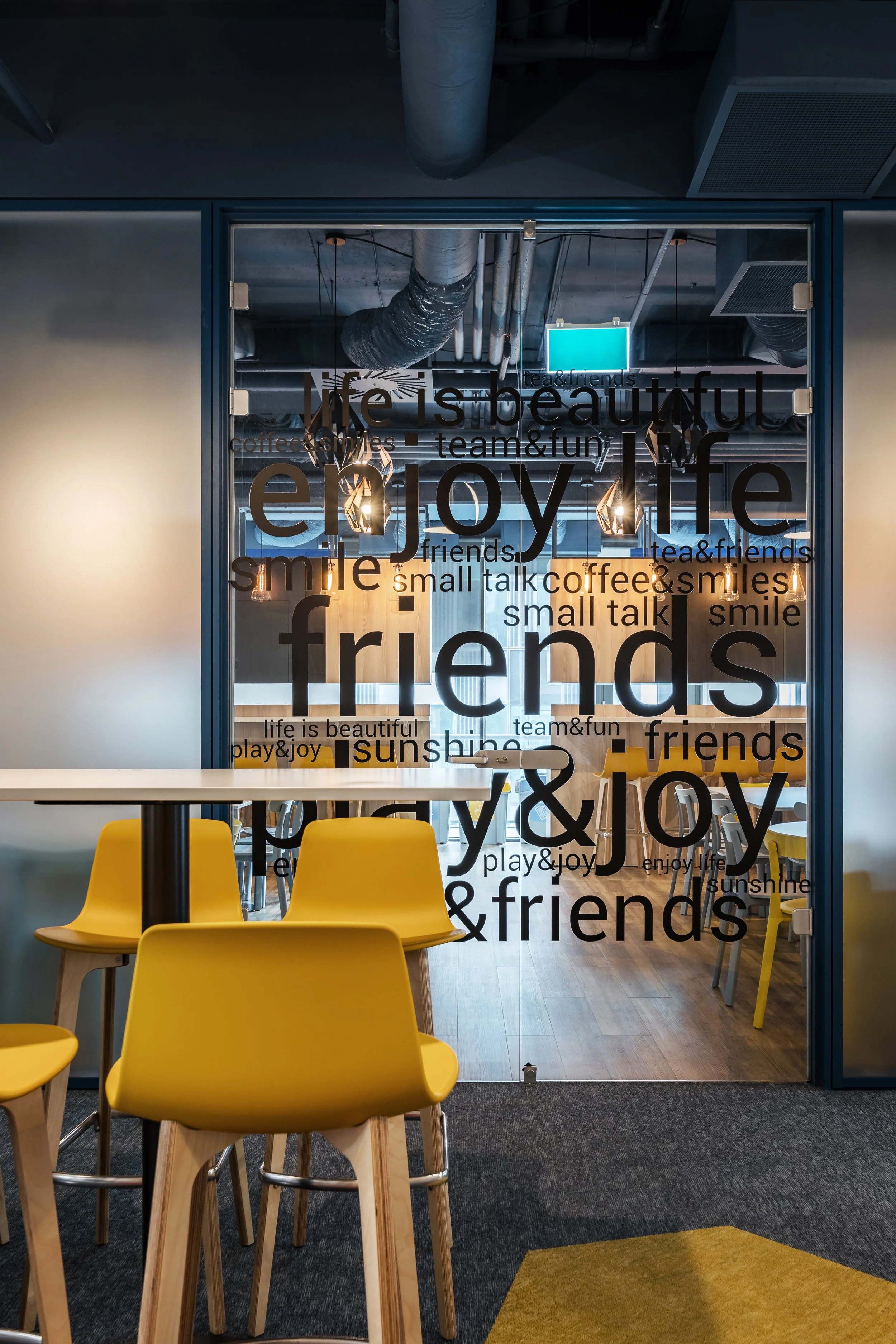 A glass door with motivational words and phrases related to friendship, happiness, and enjoyment written in various sizes and fonts. Inside the room, there are yellow bar stools and tables, with a modern, industrial-style ceiling with exposed ducts and hanging lamps.