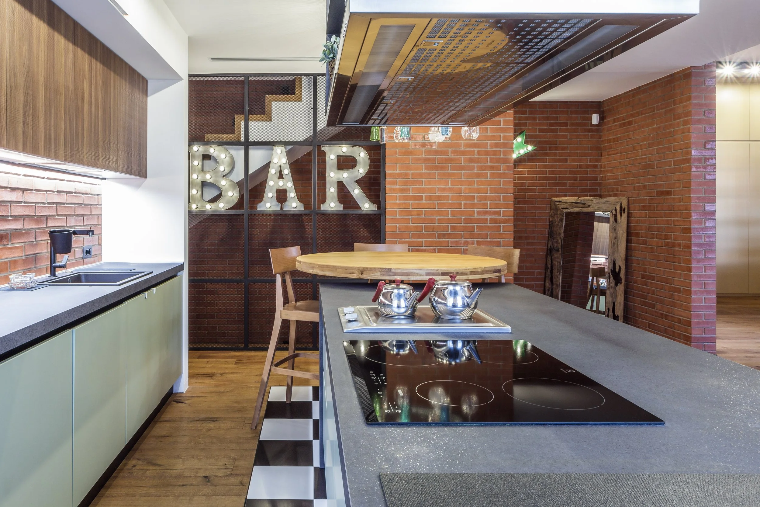 Modern kitchen with a brick wall, a black and white checkered floor, a countertop with a black induction cooktop, and a wooden round table with chairs in the background. Large illuminated