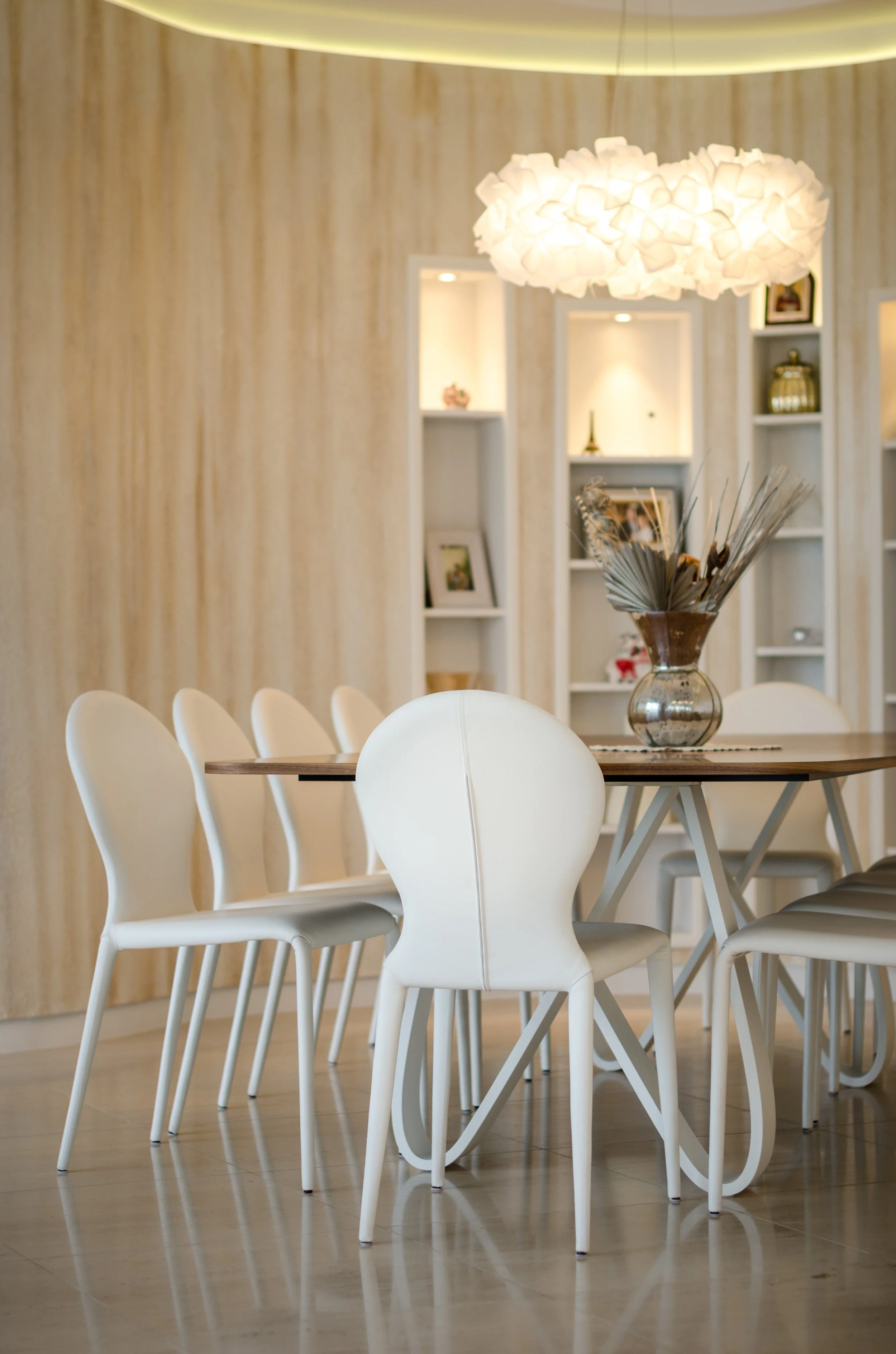 Modern dining room with white chairs around a wooden table, a large decorative white pendant light, and built-in shelves with decorative items in the background.