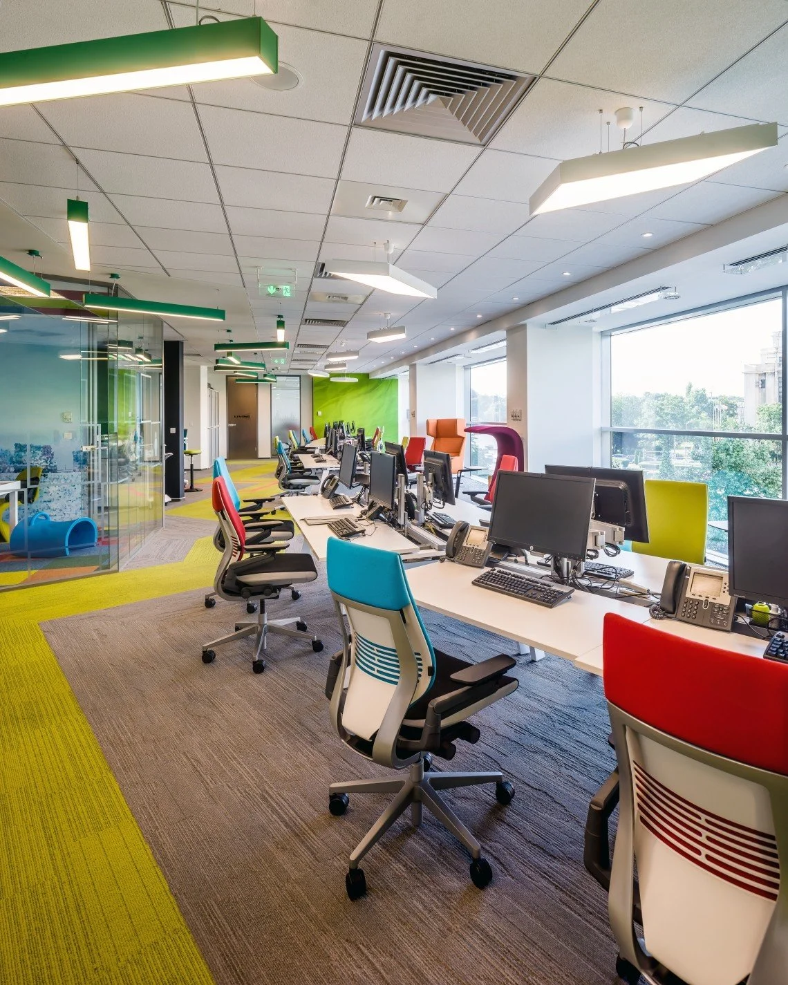 Modern, open-plan office with colorful chairs, multiple desks with computer workstations, large windows, and ceiling lighting, with a glass-walled play area in the background.