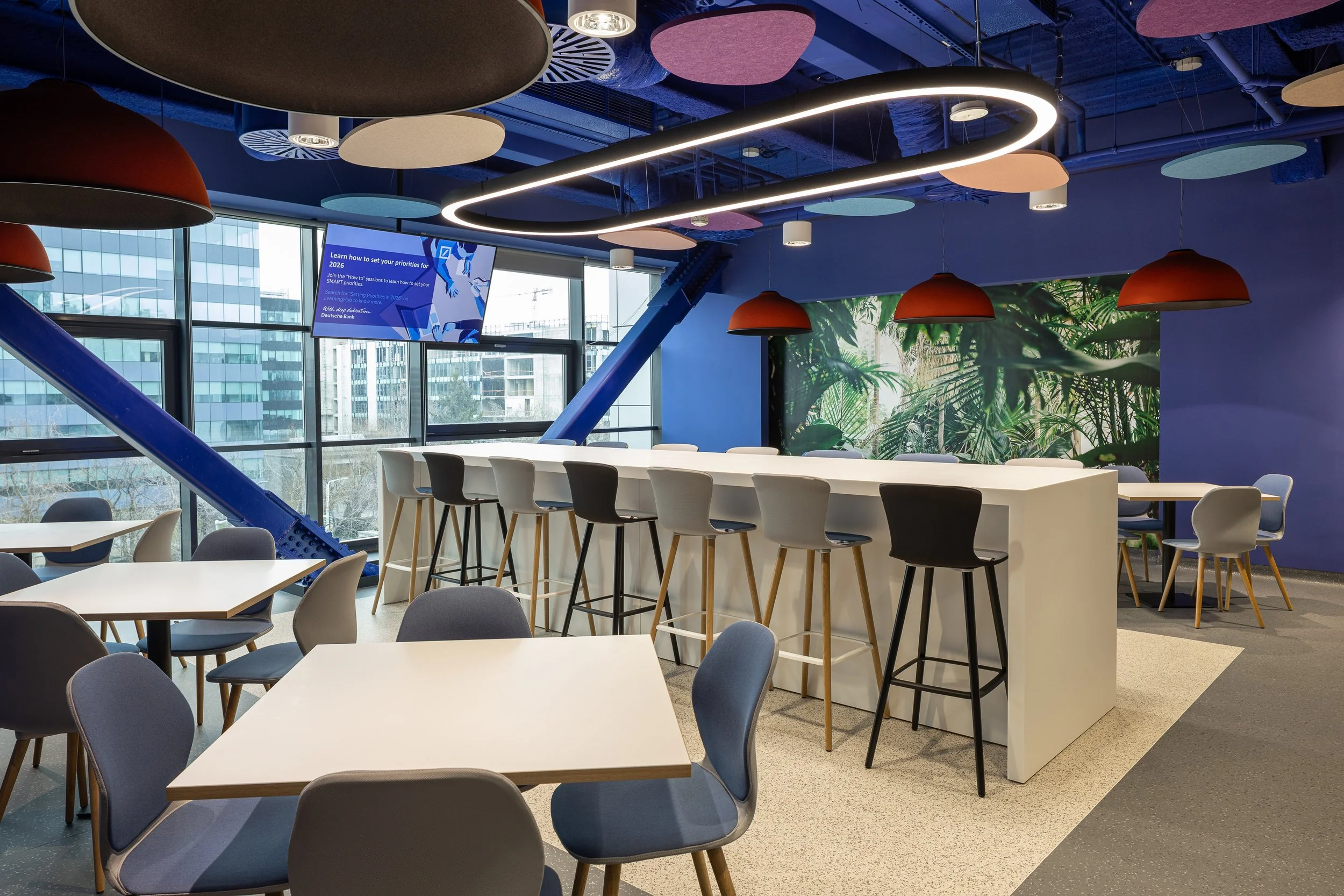 Modern office cafeteria with white tables and multicolored chairs, high bar seating with stools, a large wall mural of jungle foliage, and hanging pendant lights, overlooking city buildings through large windows.