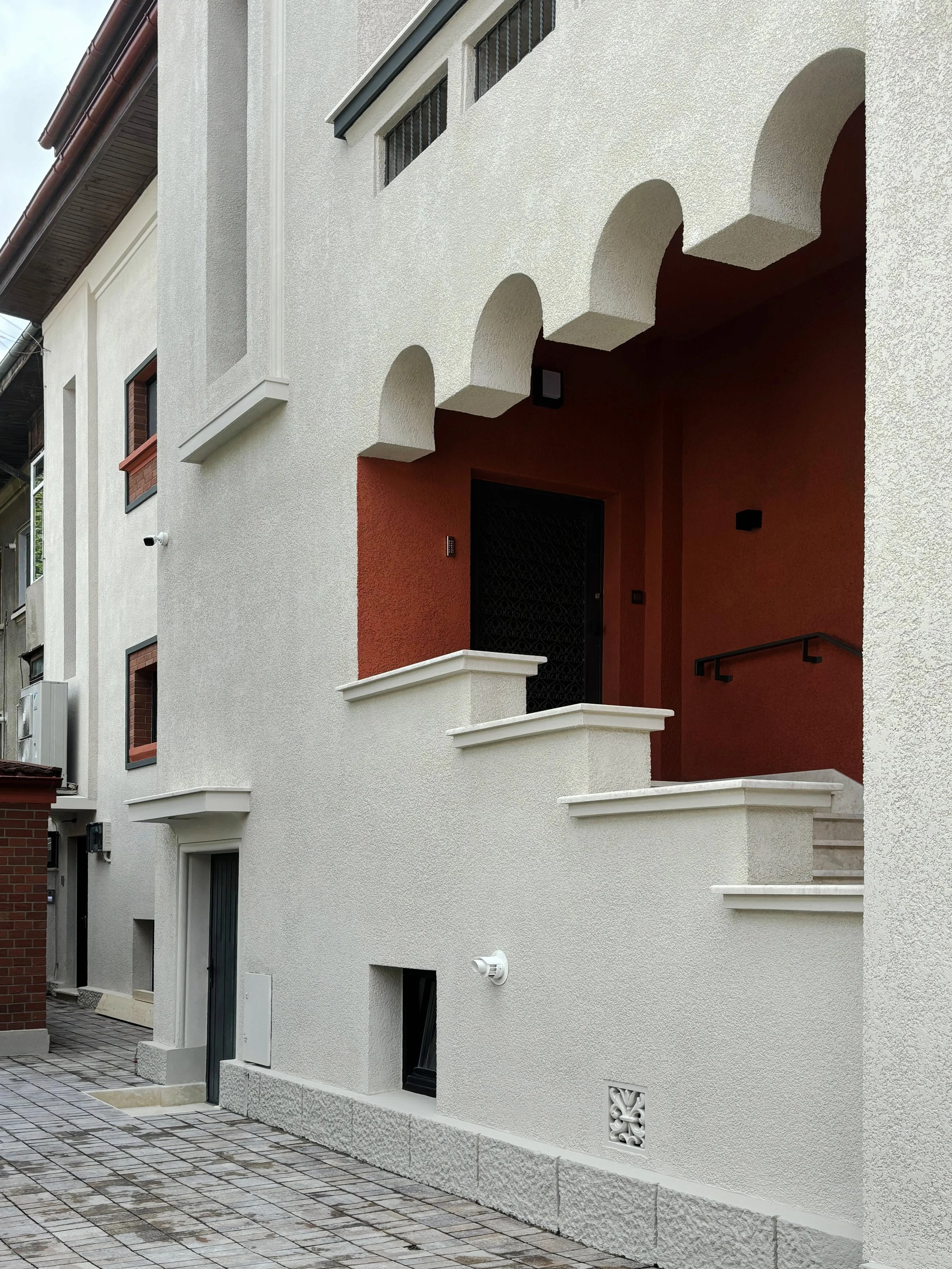 Modern residential building with arched entryway, red accent wall, and textured white exterior walls. Stone pathway in front.