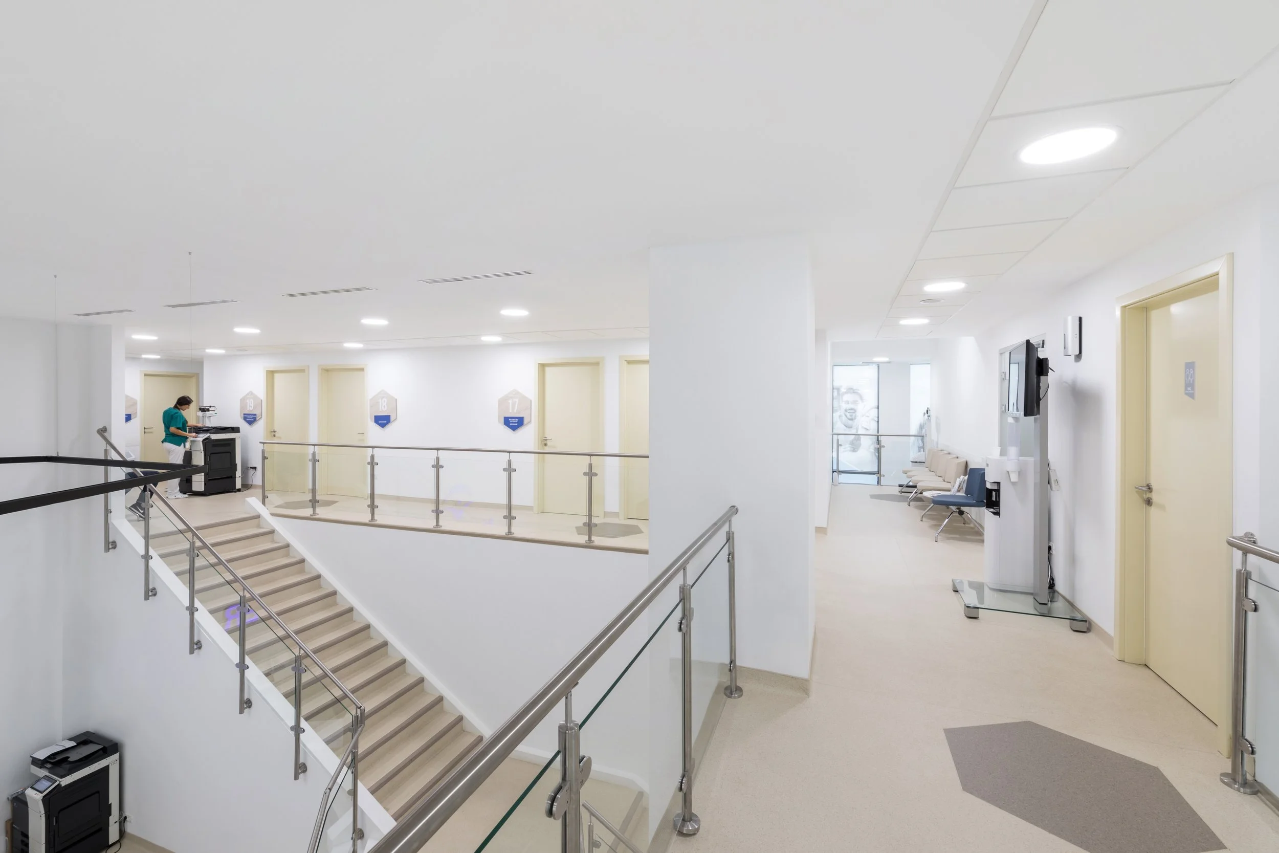 Interior view of a modern hospital corridor and reception area, featuring a staircase, seating, and medical equipment.