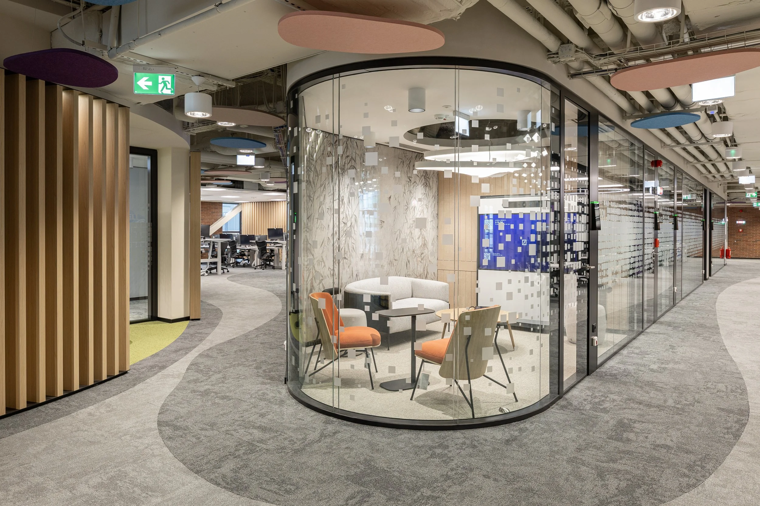 Modern office space with a glass-walled meeting room or lounge area furnished with seating and tables, and an open workspace with desks and chairs in the background.