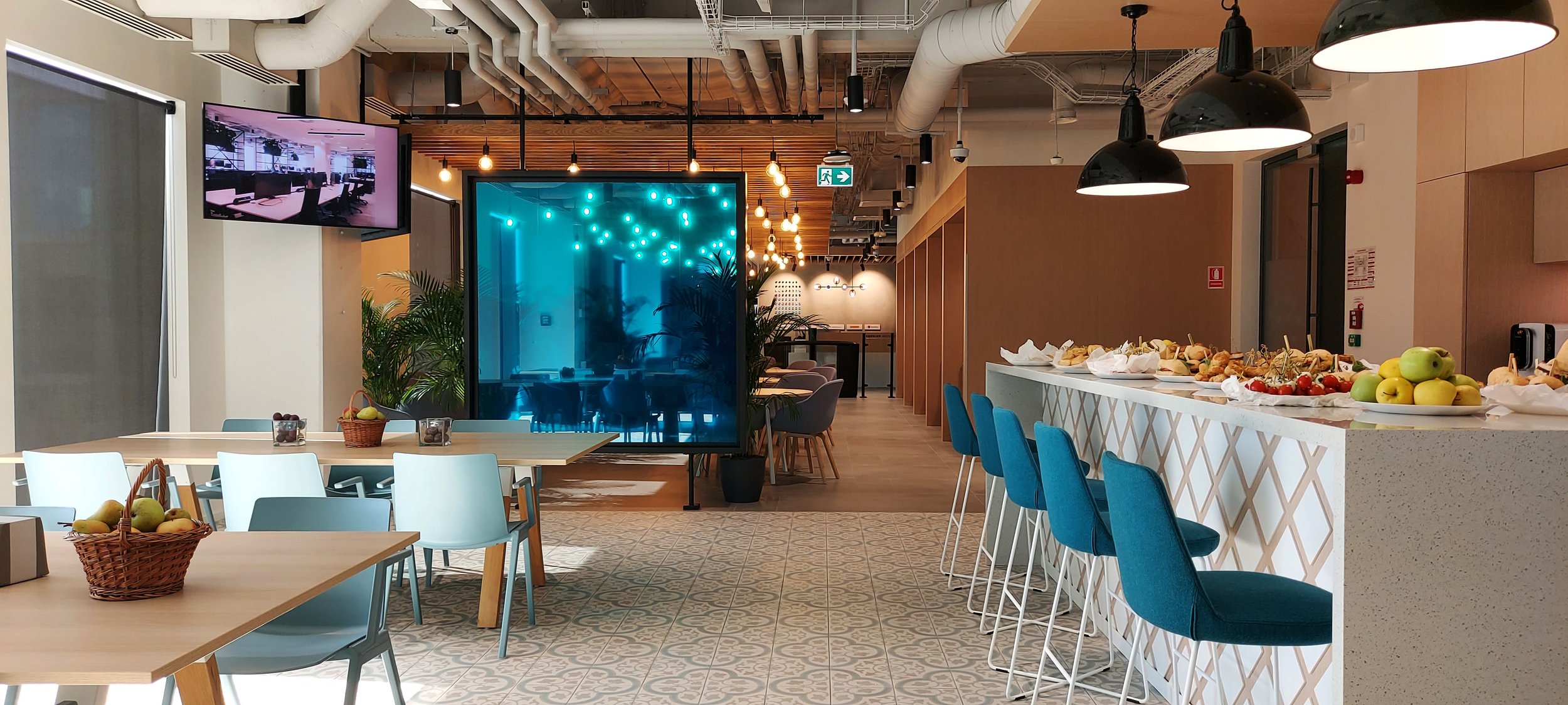 Modern cafeteria or lounge area with seating, a bar counter with fruit and appetizers, decorative lighting, plants, and a large screen display.