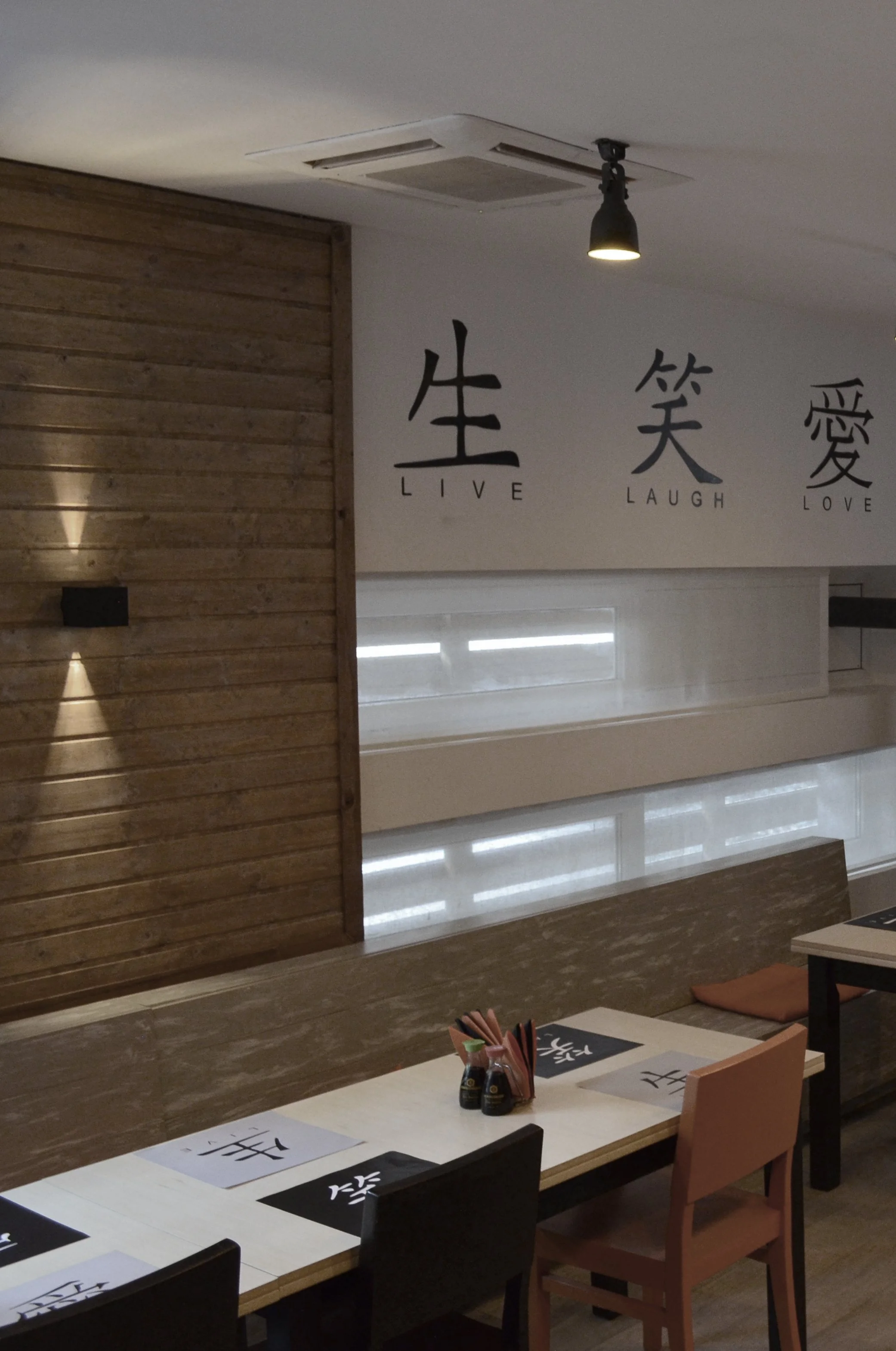 Interior of a restaurant with a wall featuring large Chinese characters and the words 'Live, Laugh, Love' underneath. There is a wooden panel and a row of tables and chairs.