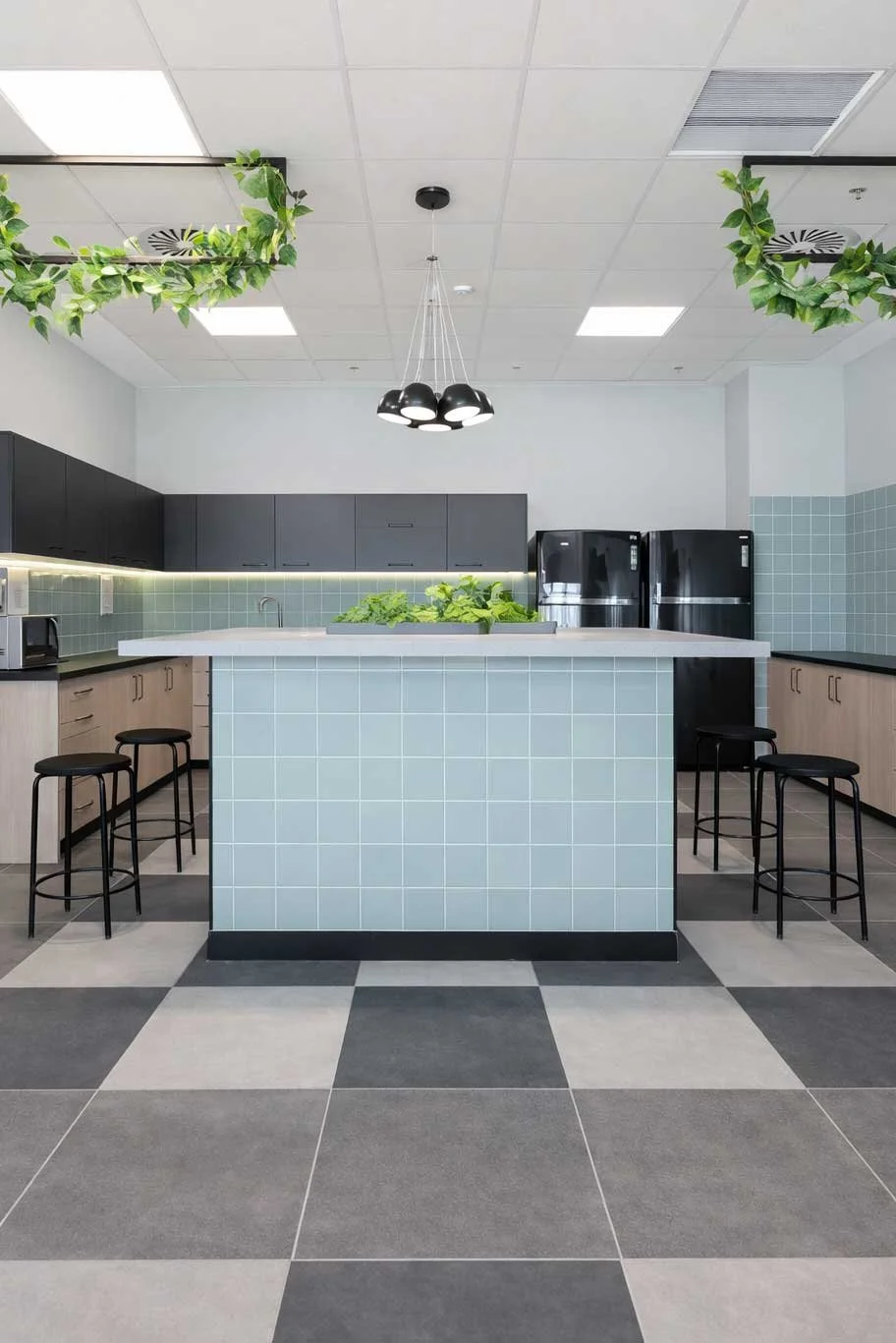 Modern kitchen with black and light wood cabinetry, black refrigerators, green tiles, and decorative green plants on the ceiling and island.