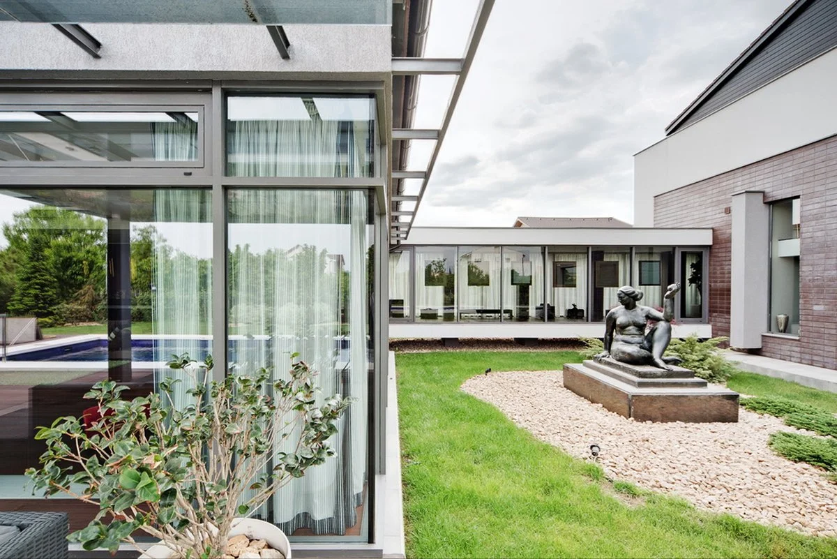 View of a modern residential backyard with glass walls, a sculpture of a seated woman, green grass, and a cloudy sky.