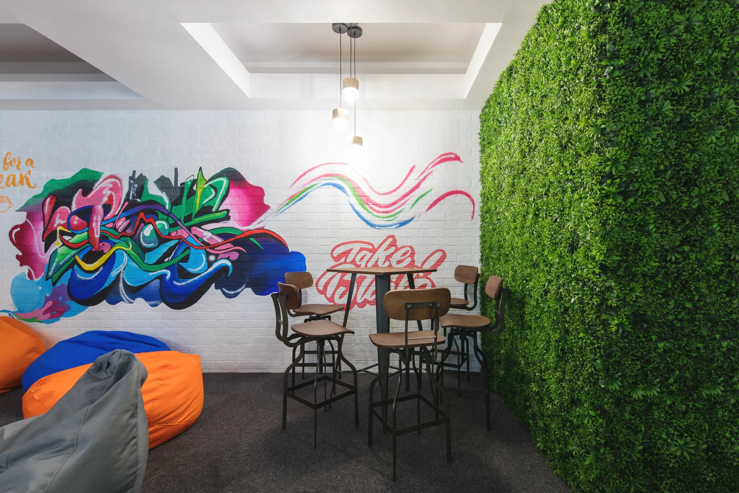 Indoor space with a white brick wall decorated with colorful graffiti art, a green wall of plants on the right, and seating including bean bag chairs and high chairs around a small table, illuminated by hanging lights.