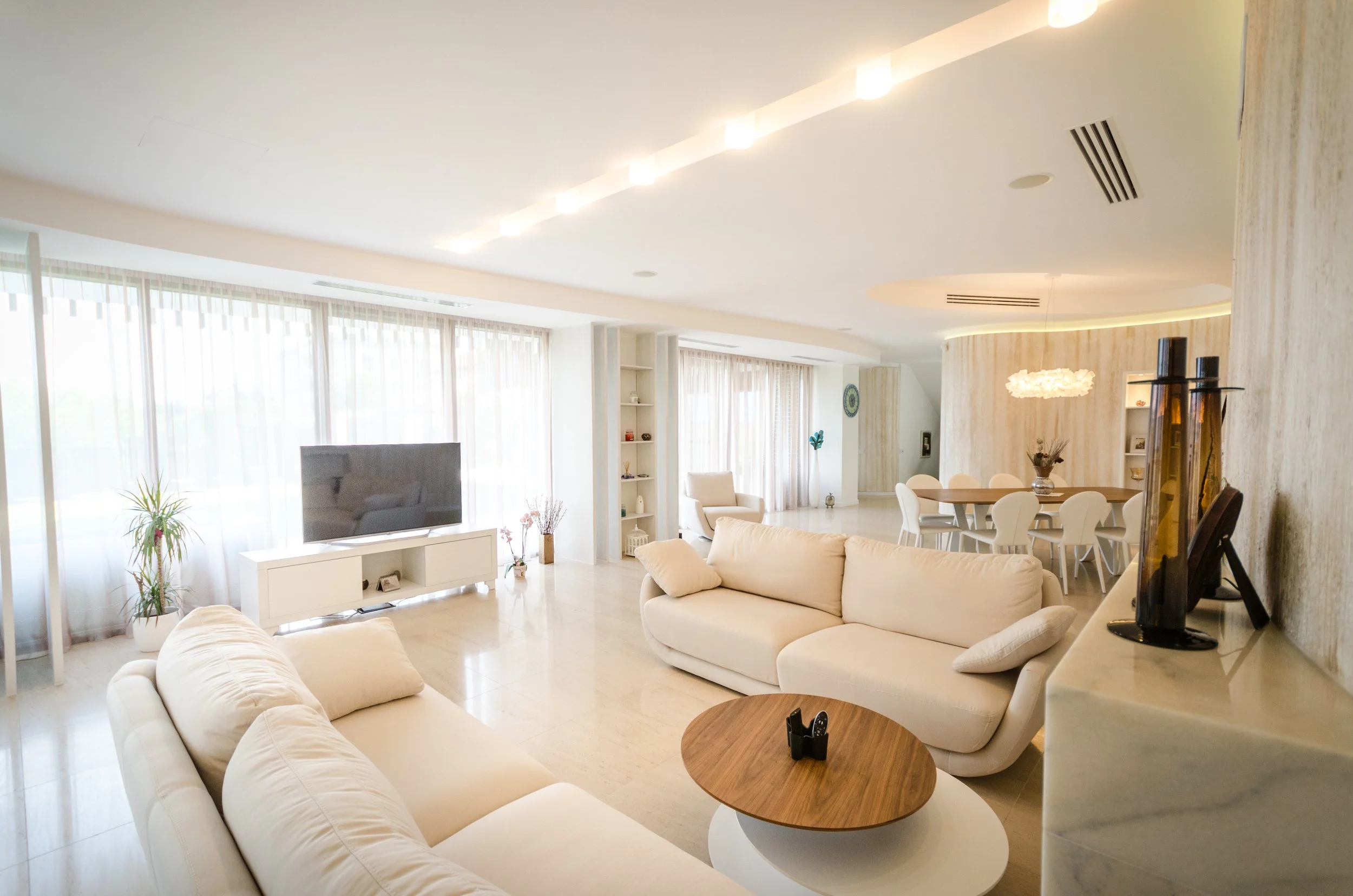 Bright, spacious living room with cream sofas, a flat-screen TV on a white console, and large windows with sheer curtains.
