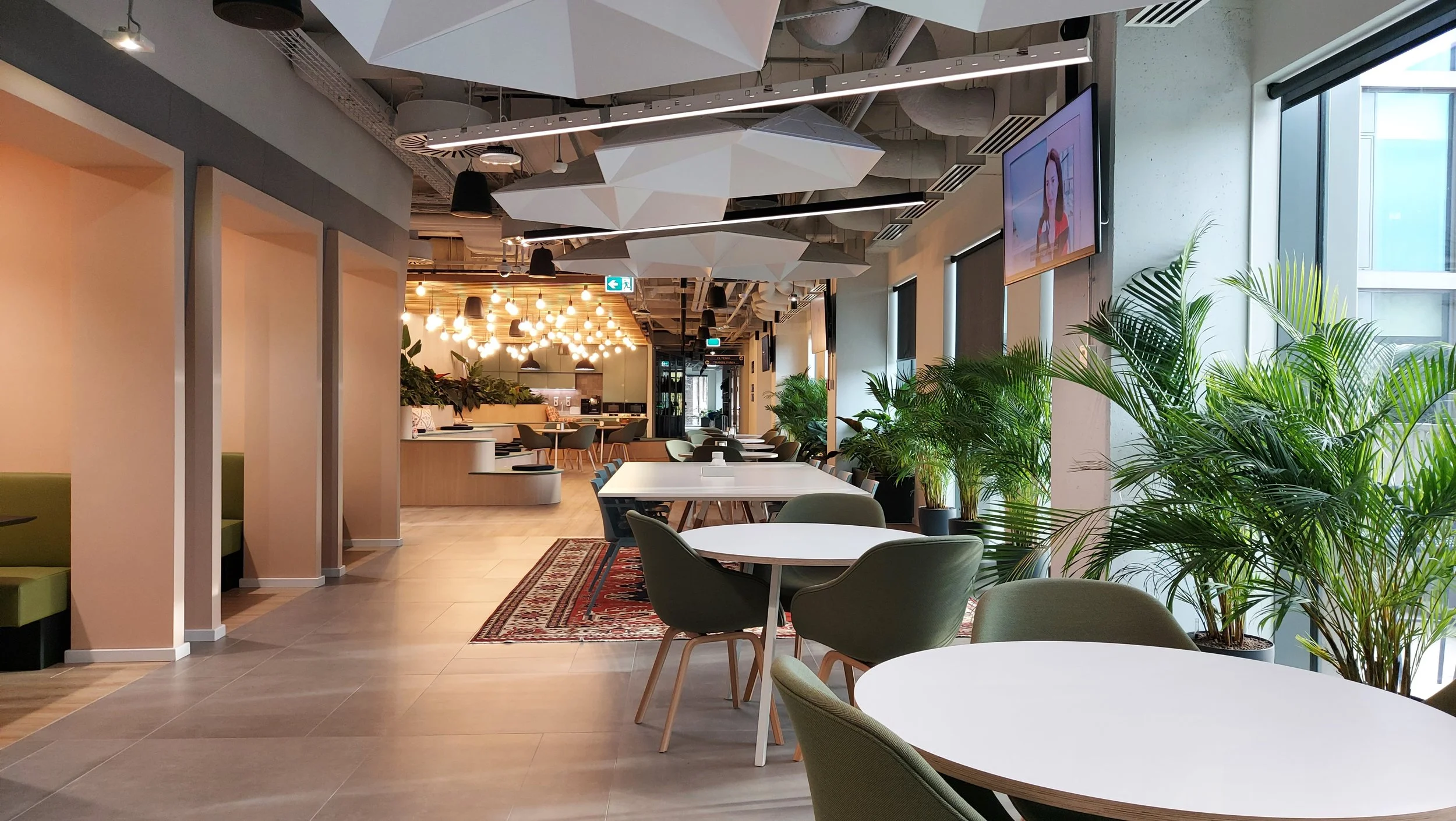 Modern office cafeteria with white tables, green chairs, large potted plants by the windows, and pendant lights.