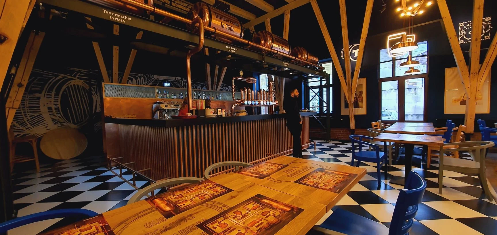 Interior of a modern bar or restaurant with a black and white checkered floor, wooden tables and chairs, a bar counter with copper pipes and taps, and artwork on dark walls illuminated by warm lighting.