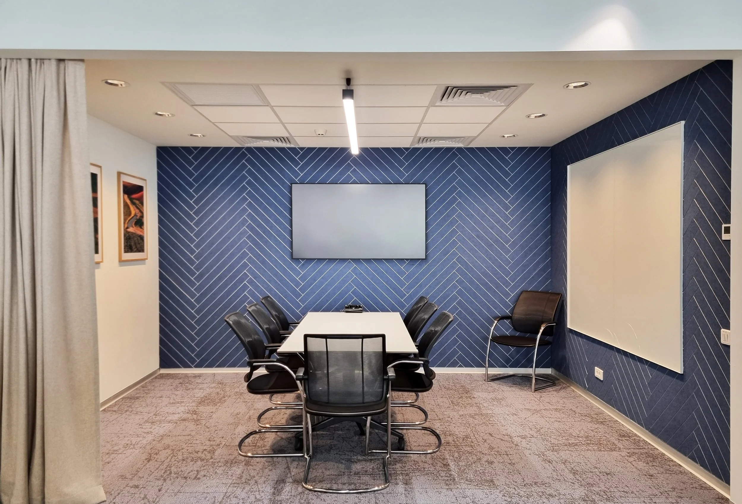 A modern conference room with a long white table, eight black office chairs, a wooden chair, a blue accent wall with white diagonal lines, a large TV screen, a whiteboard, and wall art.