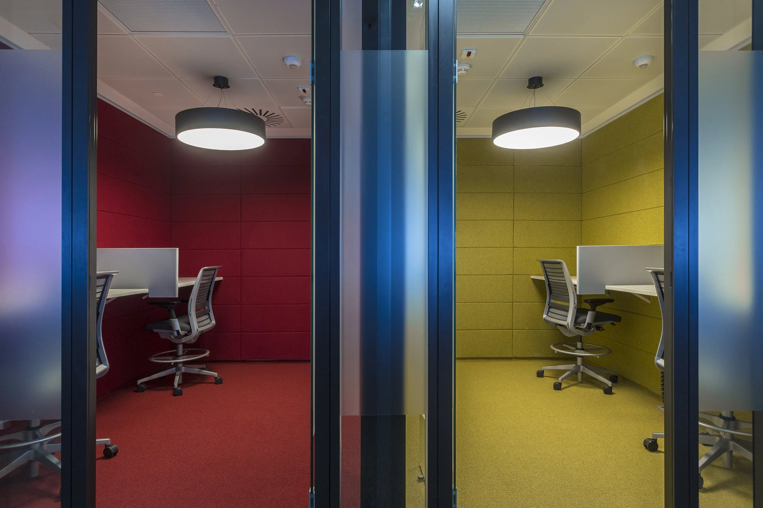 Two soundproof study booths with chairs and desks, one with red walls and the other with yellow walls, separated by a glass door.