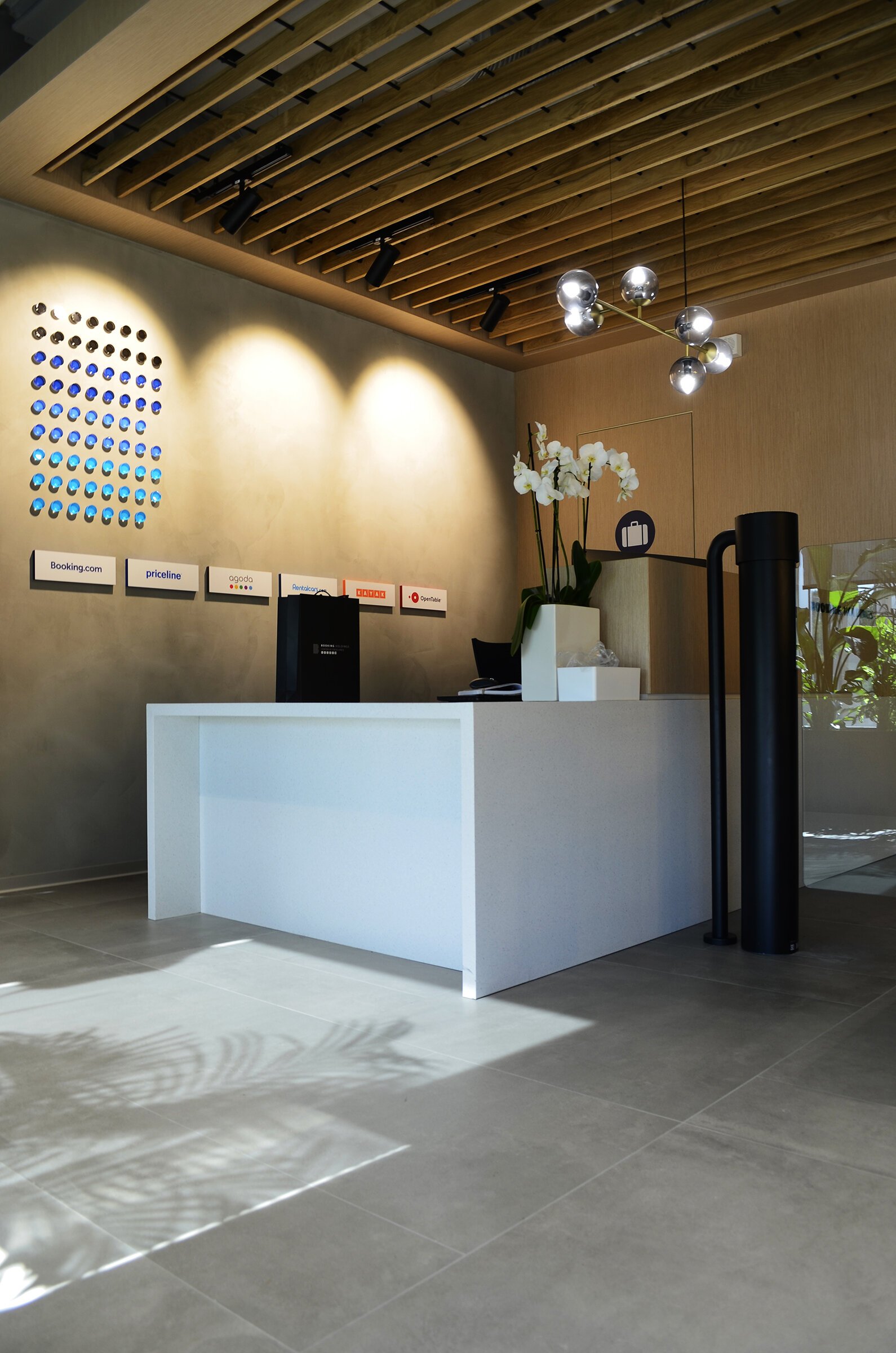 Modern hotel reception desk with a white counter, black equipment, a potted orchid, and modern lighting fixtures on a wood-paneled ceiling. Signs with popular booking and travel websites are on the wall.