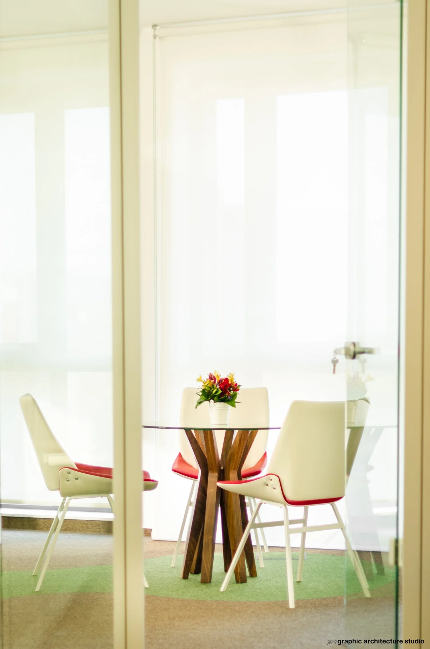 A glass round table with a wooden base, surrounded by four white chairs with red accents, in a bright room with sheer curtains, and a small vase of flowers on the table.