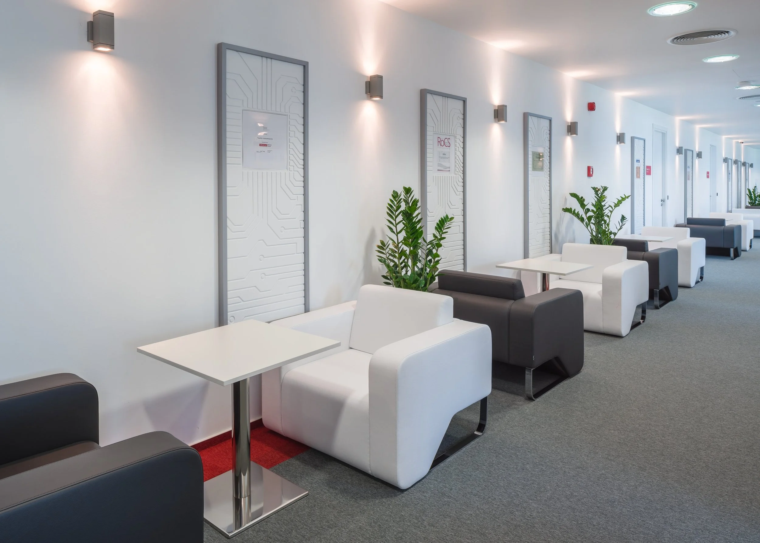 Modern office lounge with white and black armchairs, small white tables, green plants, and framed artwork on the white wall, illuminated by wall sconces.