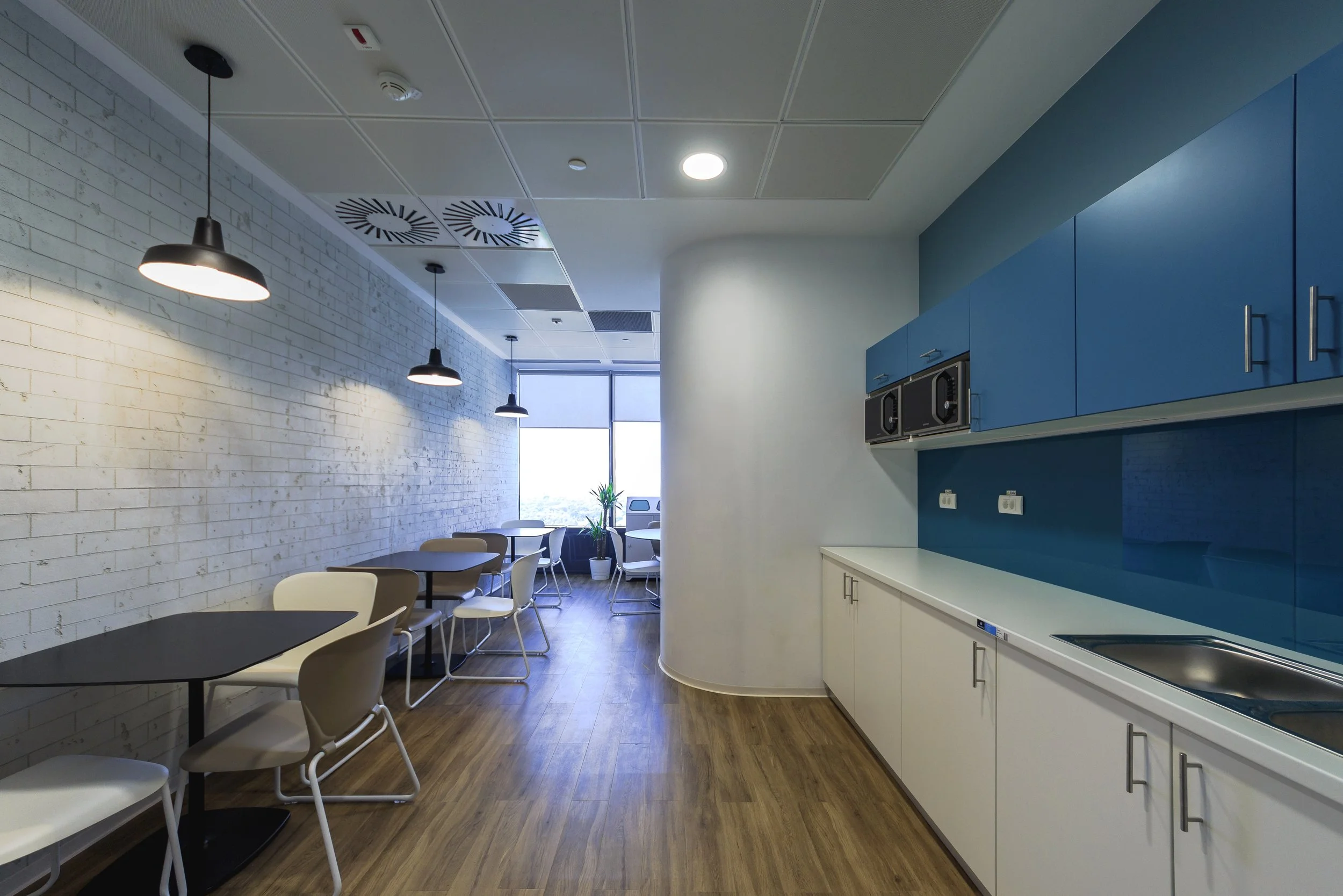 Modern office kitchenette with blue cabinets, white countertops, a sink, and two microwave ovens. Adjacent sitting area with tables, chairs, and large windows with a view.