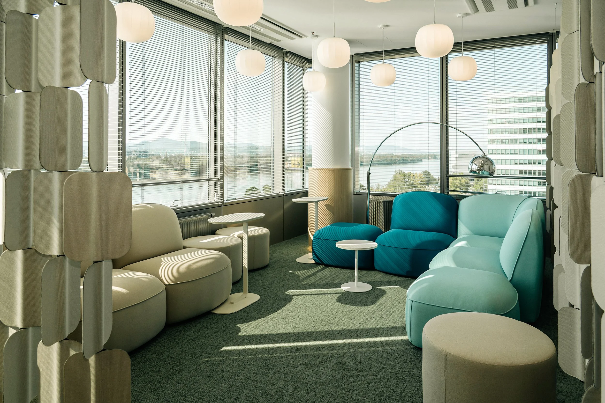 Modern lounge area with colorful curved couches, white tables, and floor-to-ceiling windows with blinds showing cityscape and water view, decorated with hanging round paper lanterns.