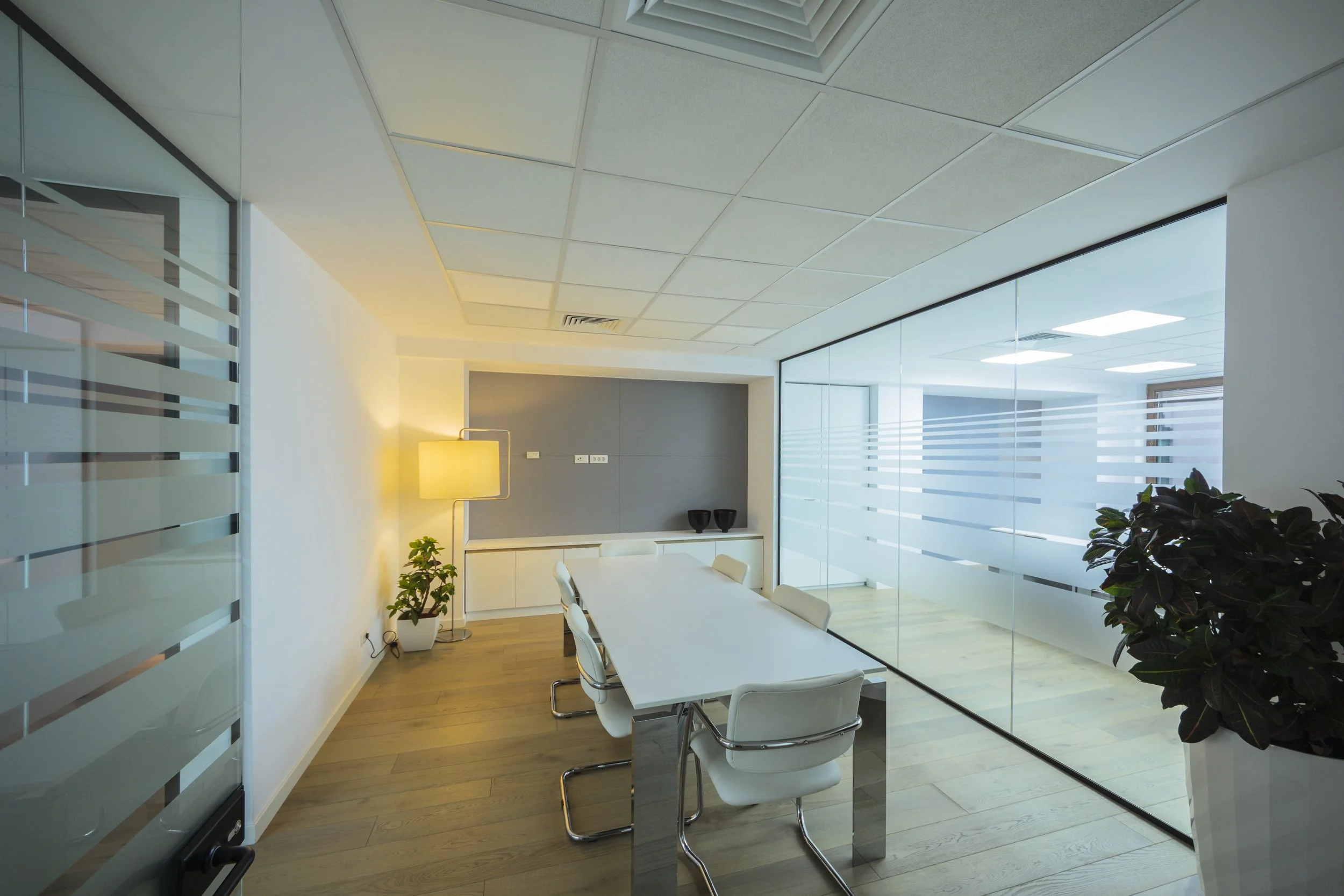 Modern office conference room with white table, white chairs, glass walls, potted plants, and a floor lamp.