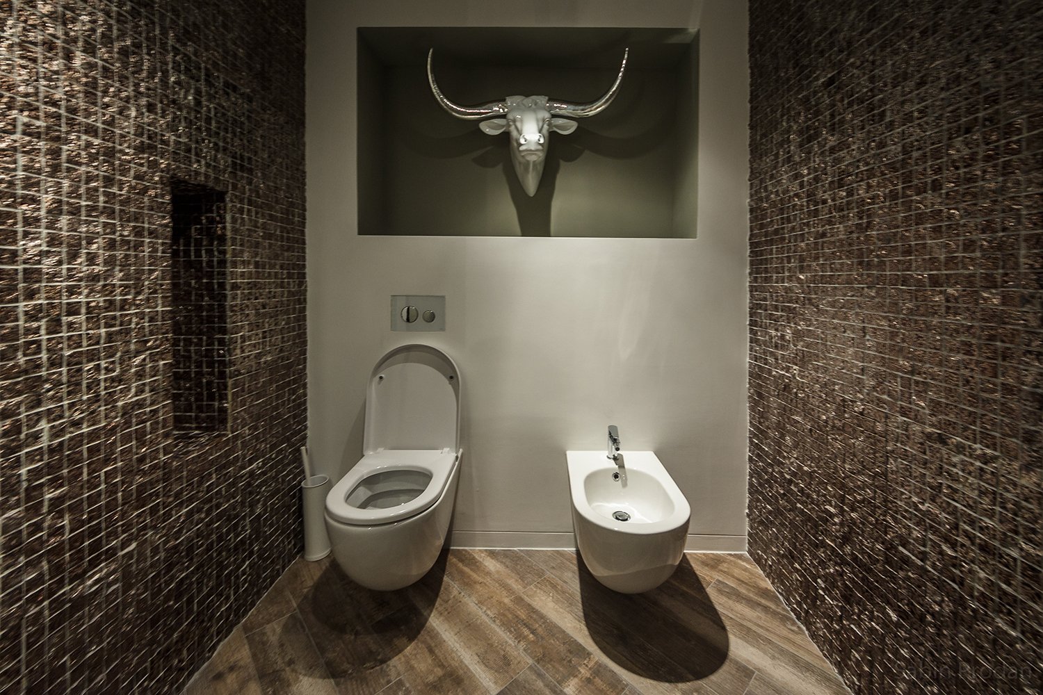 Modern bathroom with dark textured tiled walls, a toilet, a bidet, and a decorative bull's head mounted on the wall above.