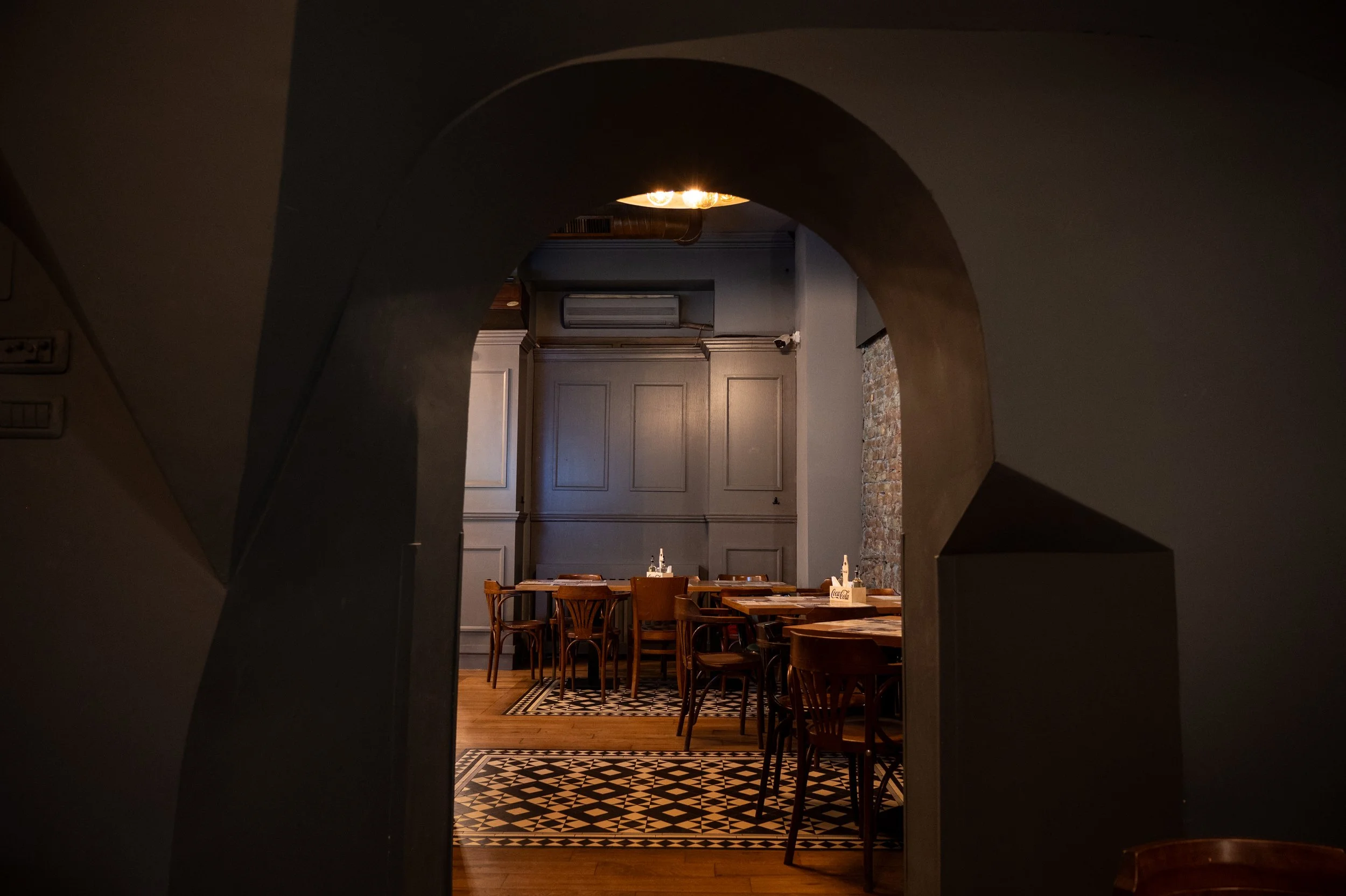 A cozy restaurant dining area seen through an arched opening. It features wooden tables and chairs, a patterned floor, gray paneled walls, and an exposed brick accent wall. Soft lighting illuminates the space.