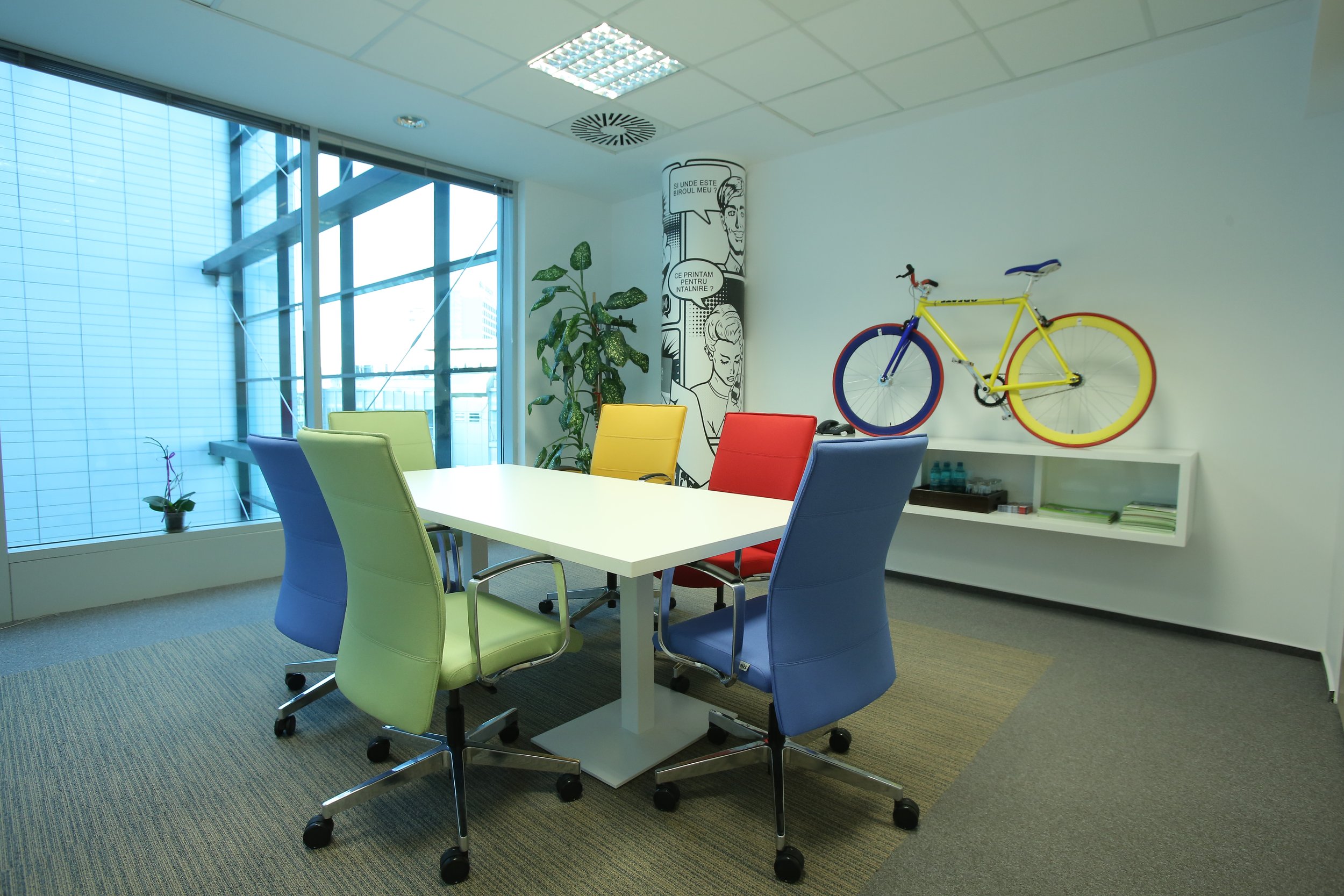 Modern office meeting room with colorful chairs around a white table, a large window with city view, a plant, a wall poster, and a yellow bicycle mounted on the wall.