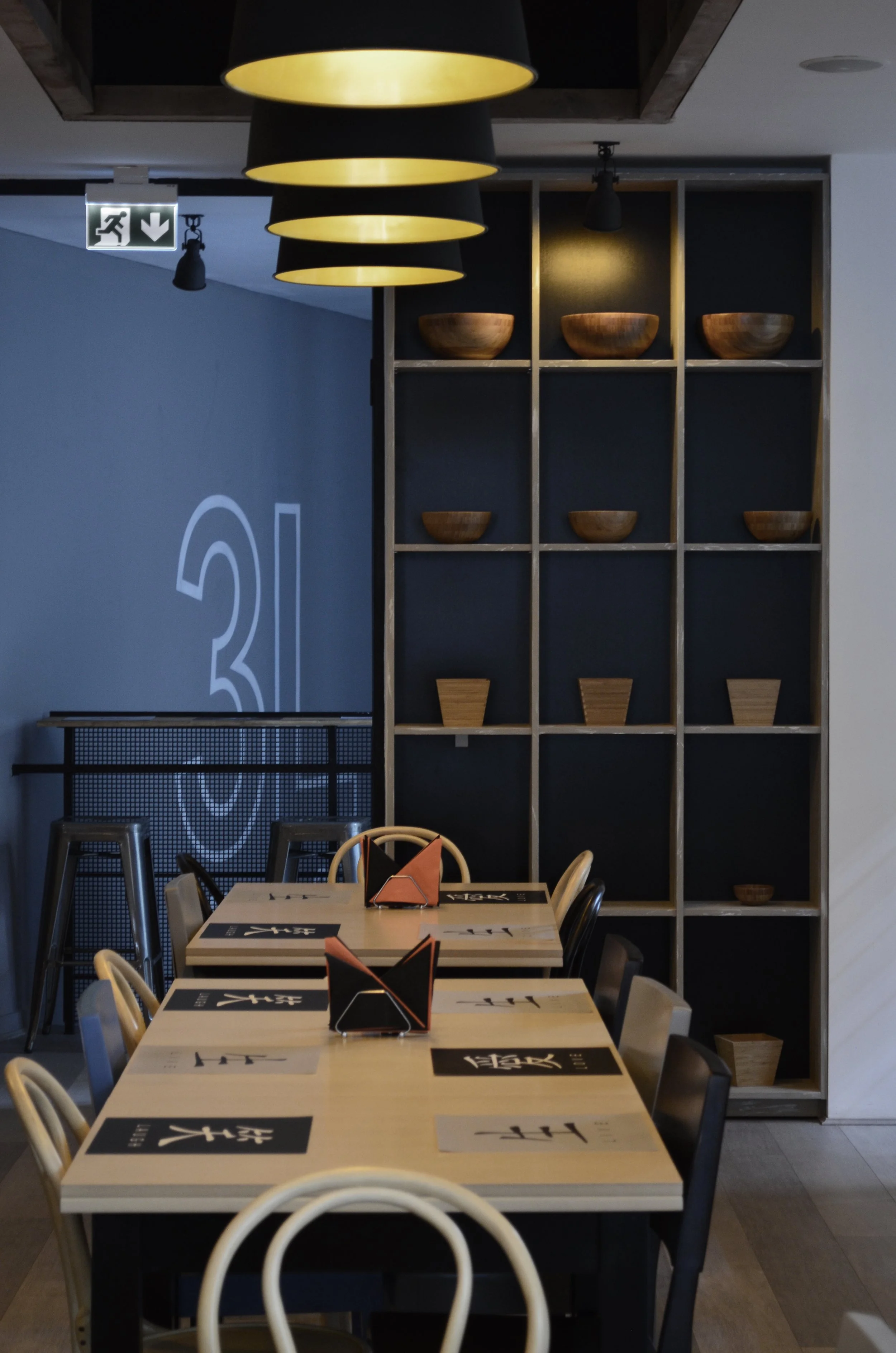 Interior of a modern restaurant with a long wooden table set for dining, black chairs, and a large black wooden shelving unit with wooden bowls. Overhead, a cluster of large black pendant lights.