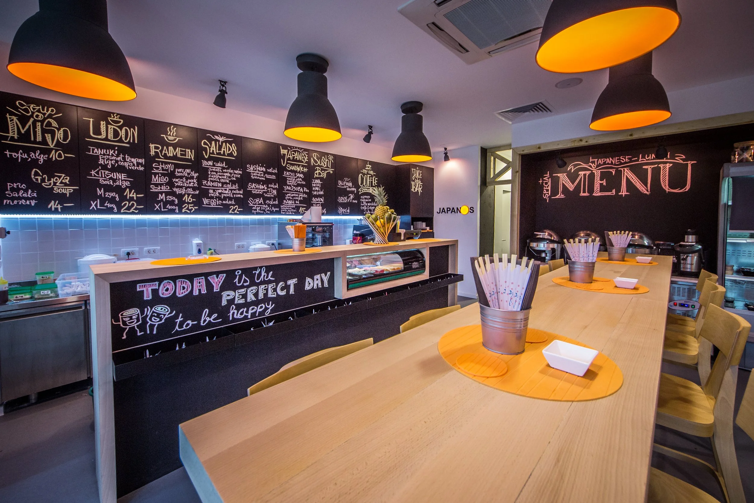 Interior of a Japanese restaurant with a long wooden table, metal pail holders with chopsticks, a black chalkboard menu, and black pendant lights with a yellow interior overhead.