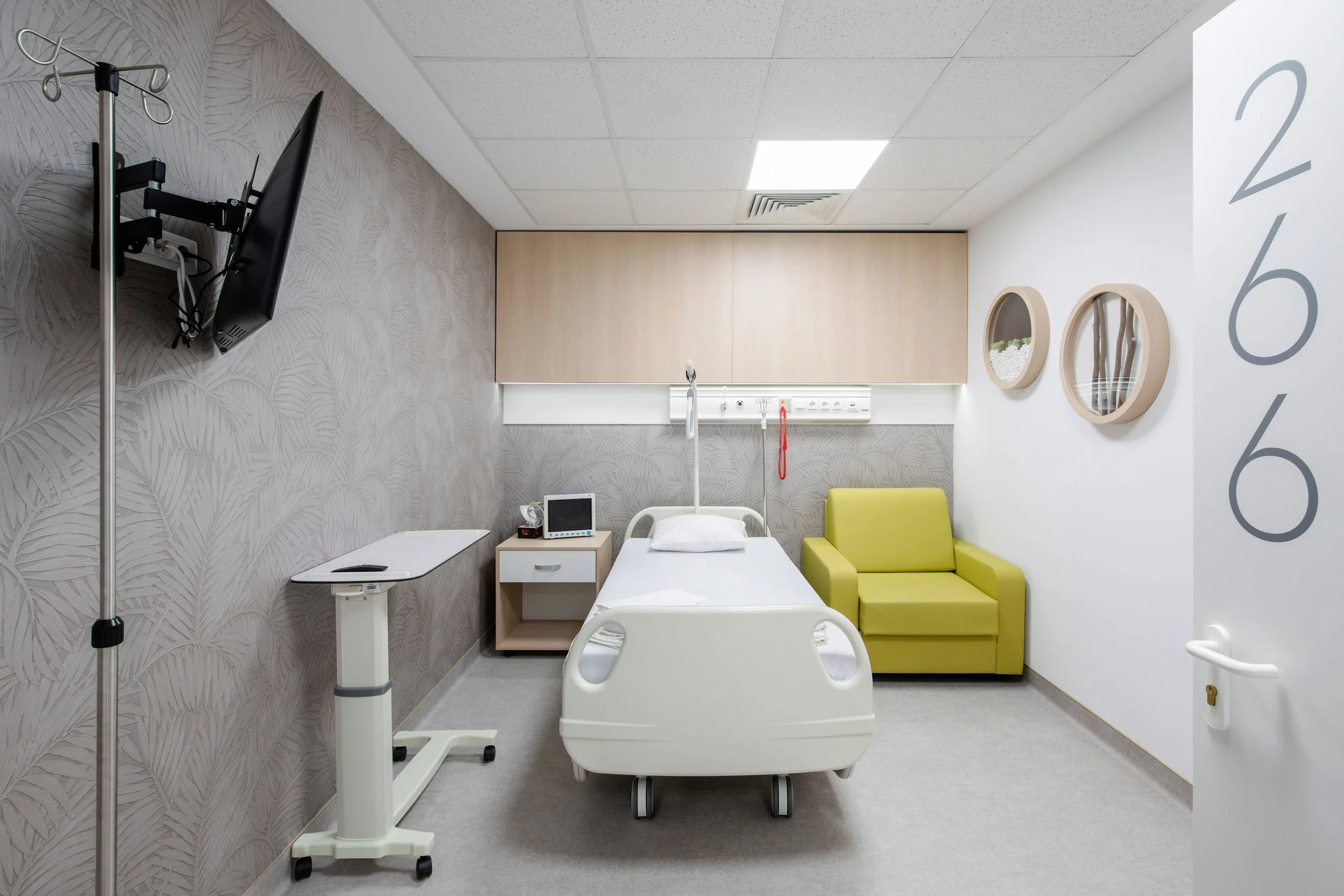 Hospital room with bed, television, and medical equipment, featuring a yellow armchair and decorative wall art.