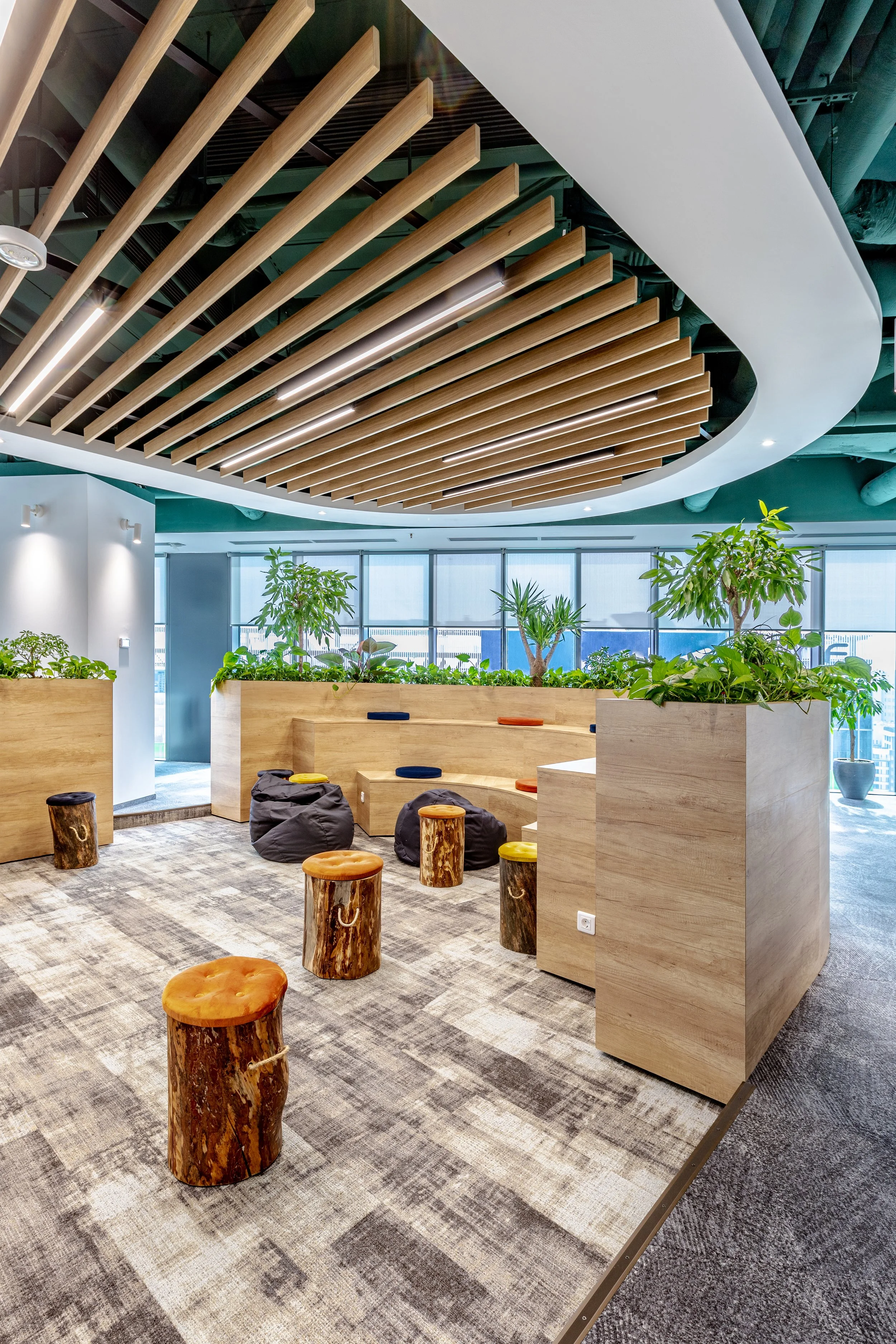 Modern office lounge with wooden stools, bean bags, and planters with green plants, under a decorative wooden ceiling design.