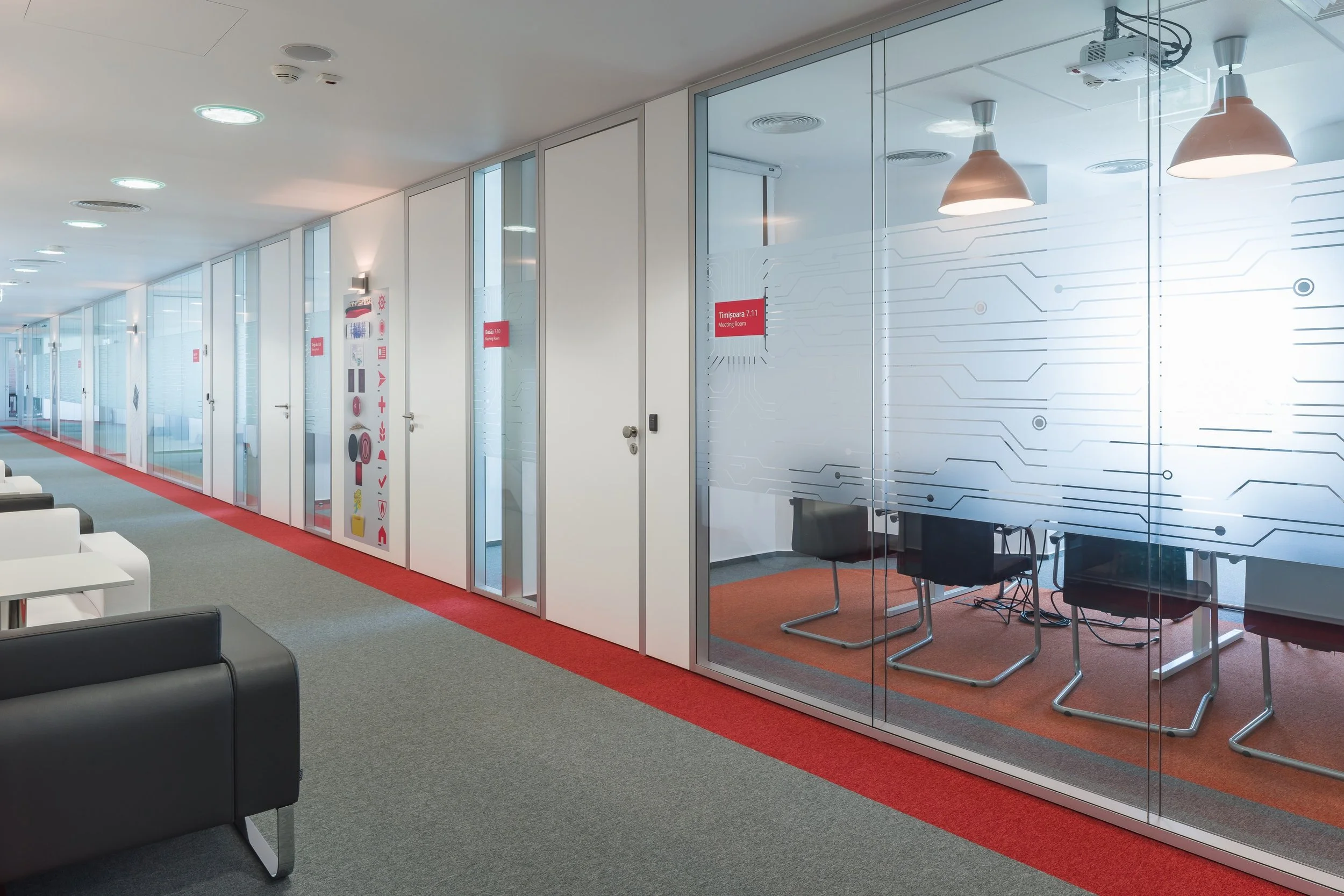 Modern office corridor with glass-walled meeting rooms, some with chairs inside, and a lounge area with white and black armchairs. The corridor has a red carpet strip along the wall and gray carpet flooring, illuminated by ceiling lights and pendant lamps.