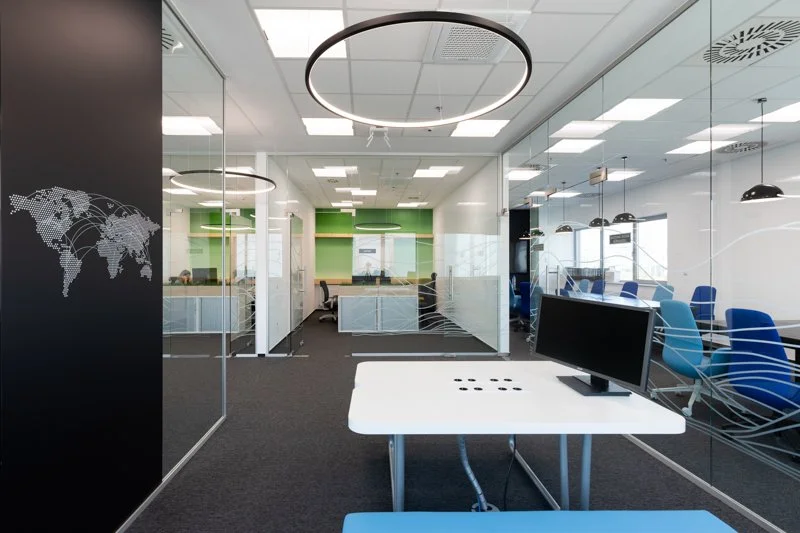 Modern office conference room with glass walls, blue chairs, a white table, and a monitor.