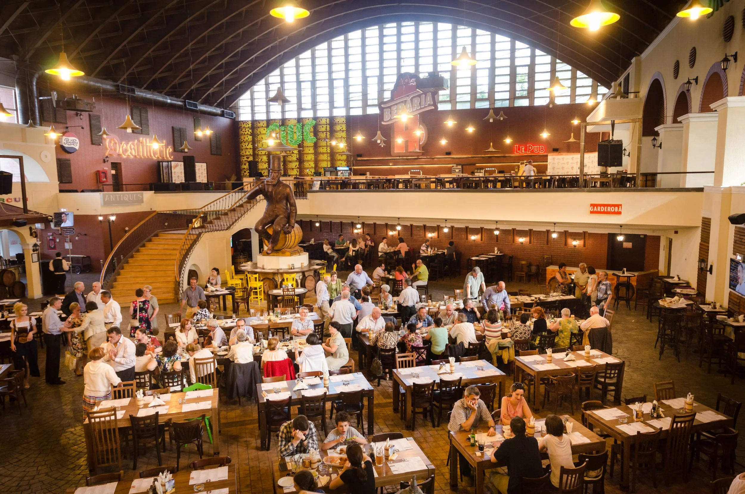 A busy restaurant with many tables filled with diners, a large bronze statue of a man sitting on a cylinder, a staircase, and a high arched ceiling with large windows and hanging yellow lights.