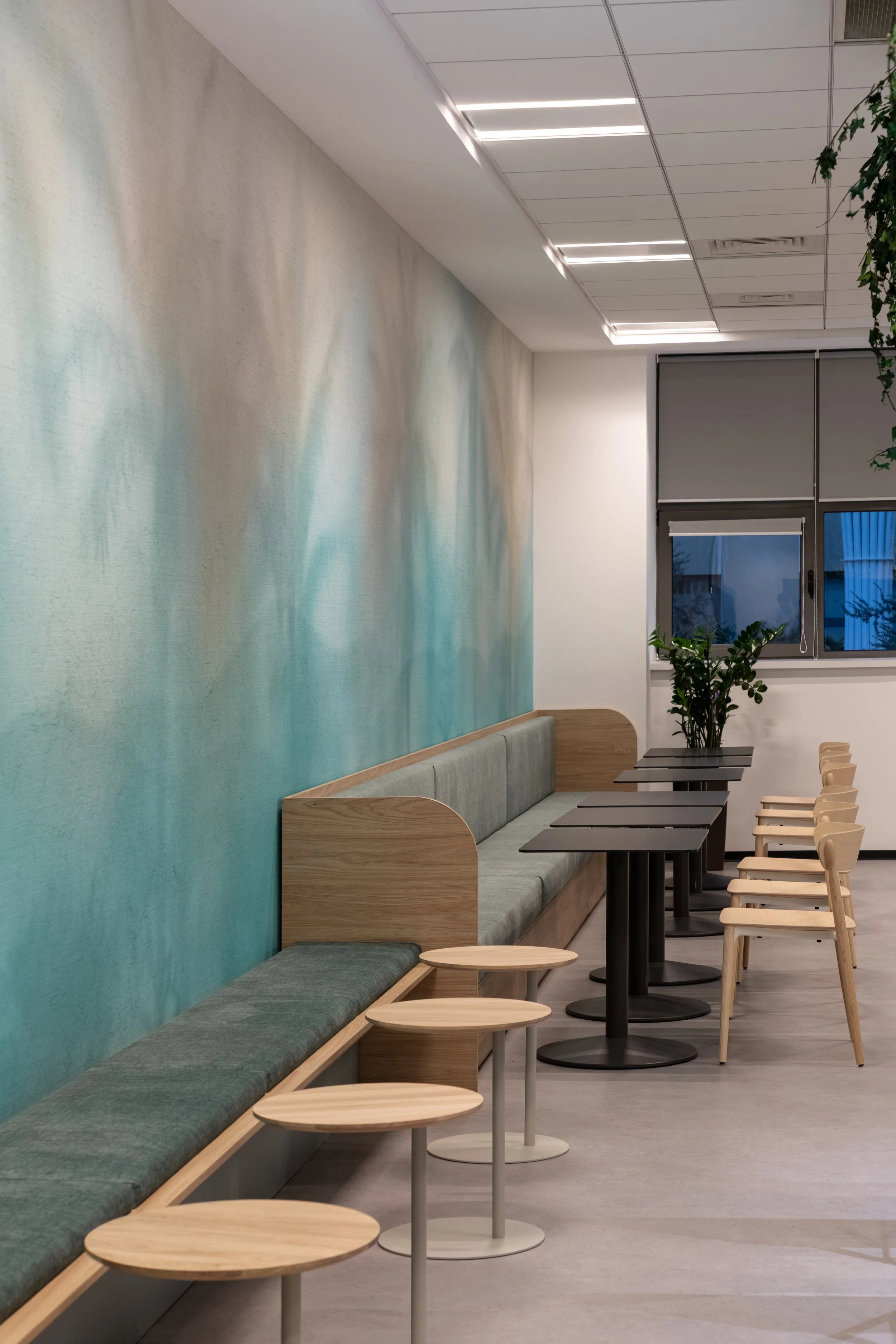 Empty modern cafe or lounge with beige and wooden furniture, a green upholstered bench along a teal gradient wall, small round tables, and wooden chairs. A window with a roller shade and potted plants are also visible.