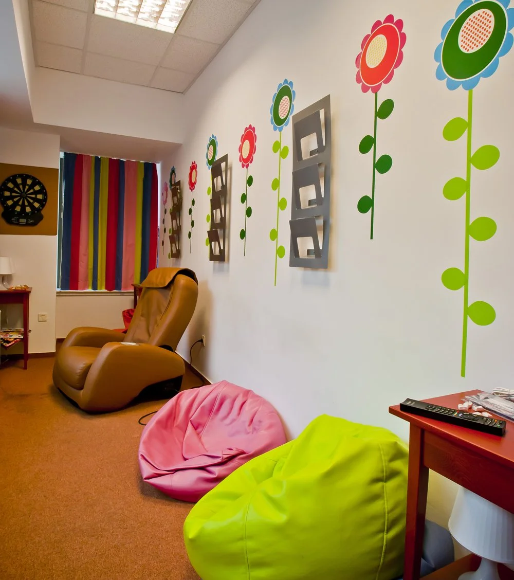 Colorful room featuring a brown massage chair, two pink and lime green bean bag chairs, layered wall decor with stylized flowers and metallic frames, a dartboard on the wall, and a window with multicolored curtains.