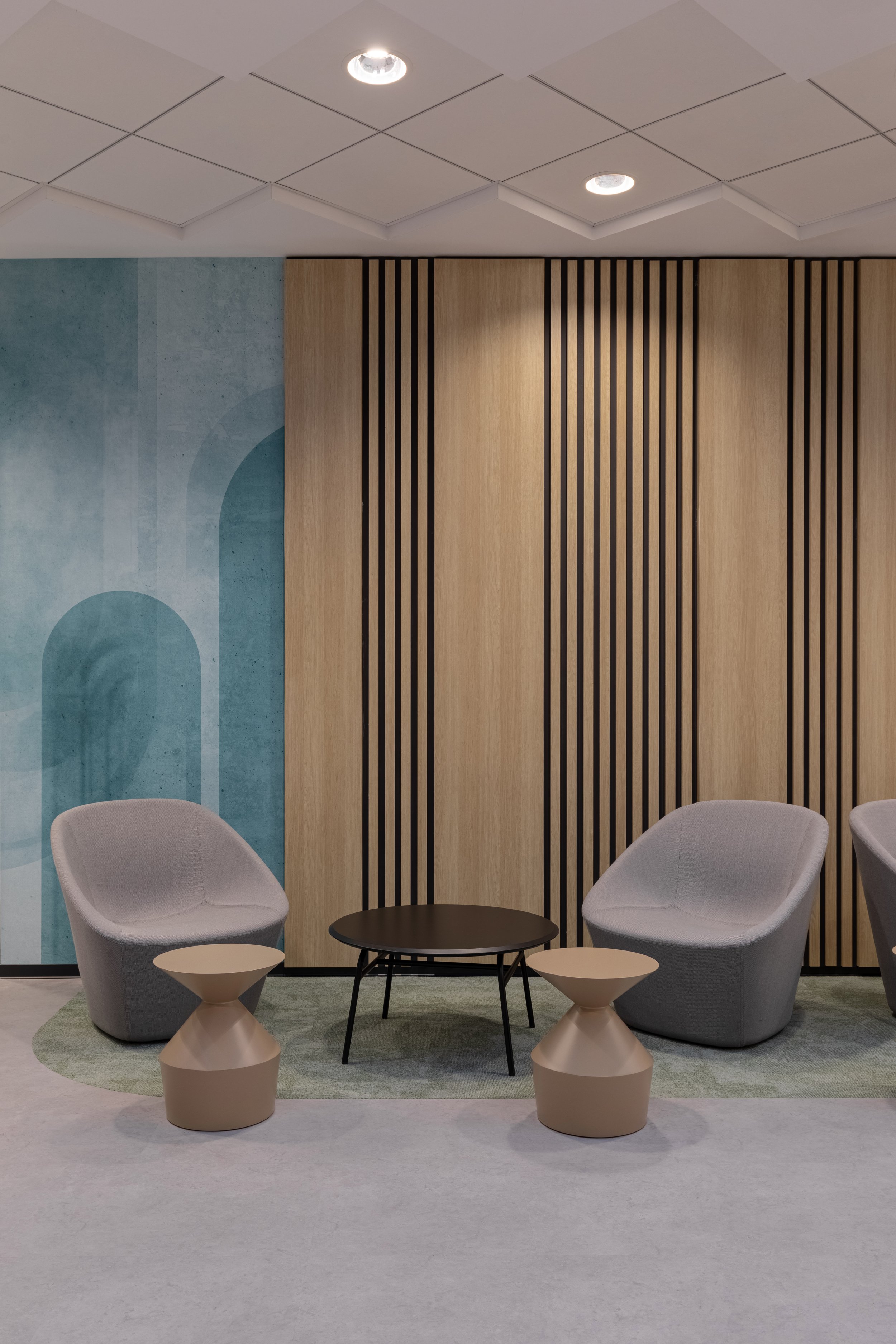 Modern office lounge with two gray armchairs, a black coffee table, and two beige side tables, set against a background of wood paneling and a blue abstract wall mural, with ceiling lights.