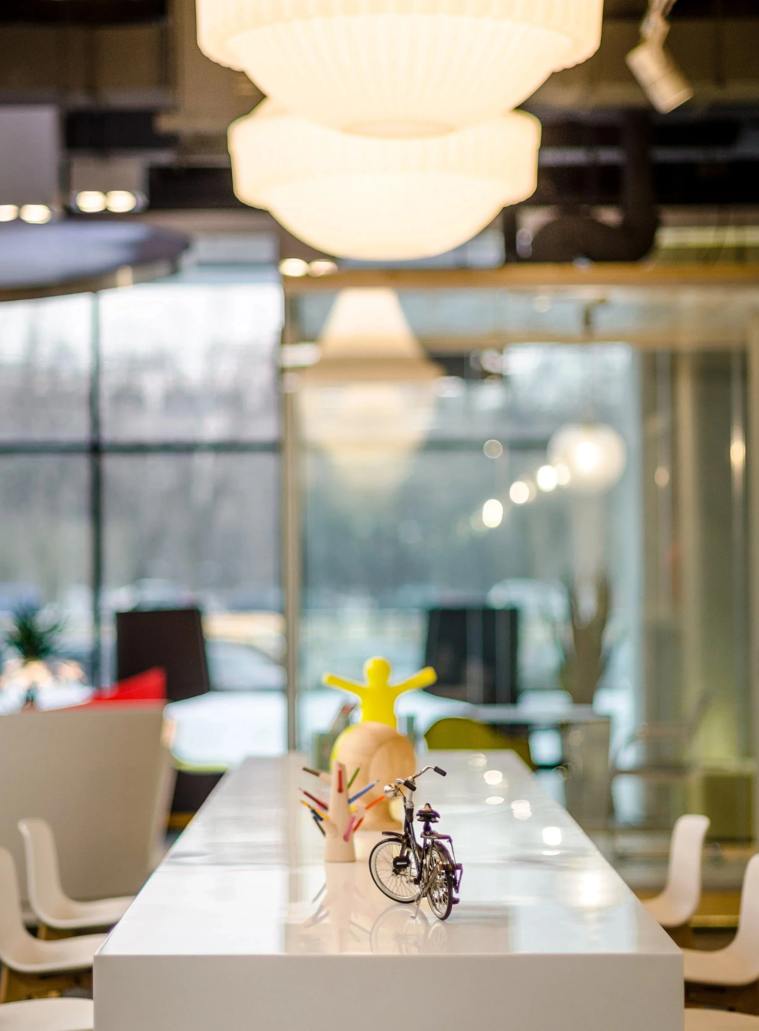 Modern indoor space with a white table decorated with miniature bicycle, colorful pens in a holder, and a yellow mannequin figure. Large, round hanging lights and floor-to-ceiling windows in the background.
