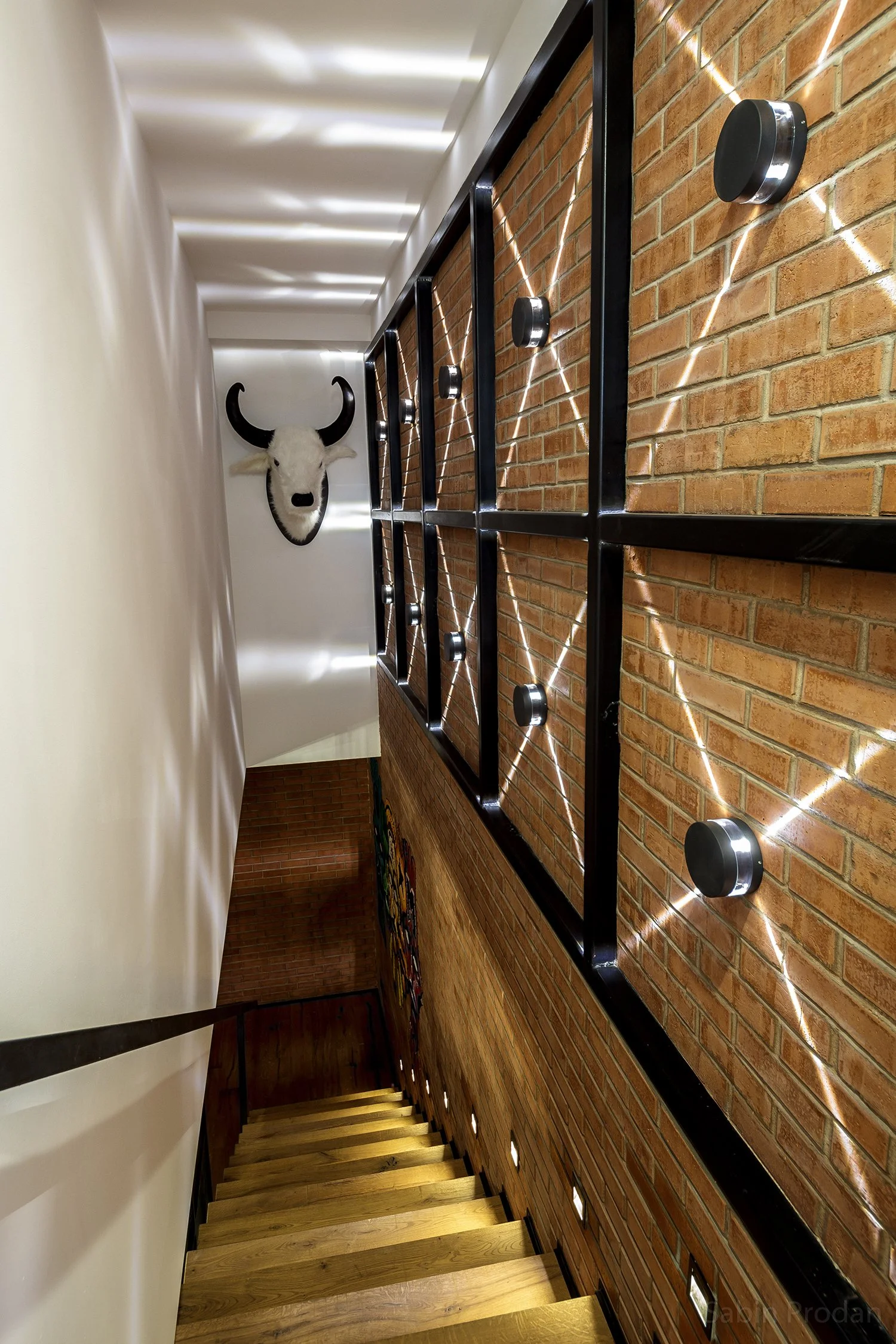 Indoor staircase with wooden steps, black metal railing, exposed brick wall with modern lighting and decorative lights, and a mounted animal skull with horns on the wall.
