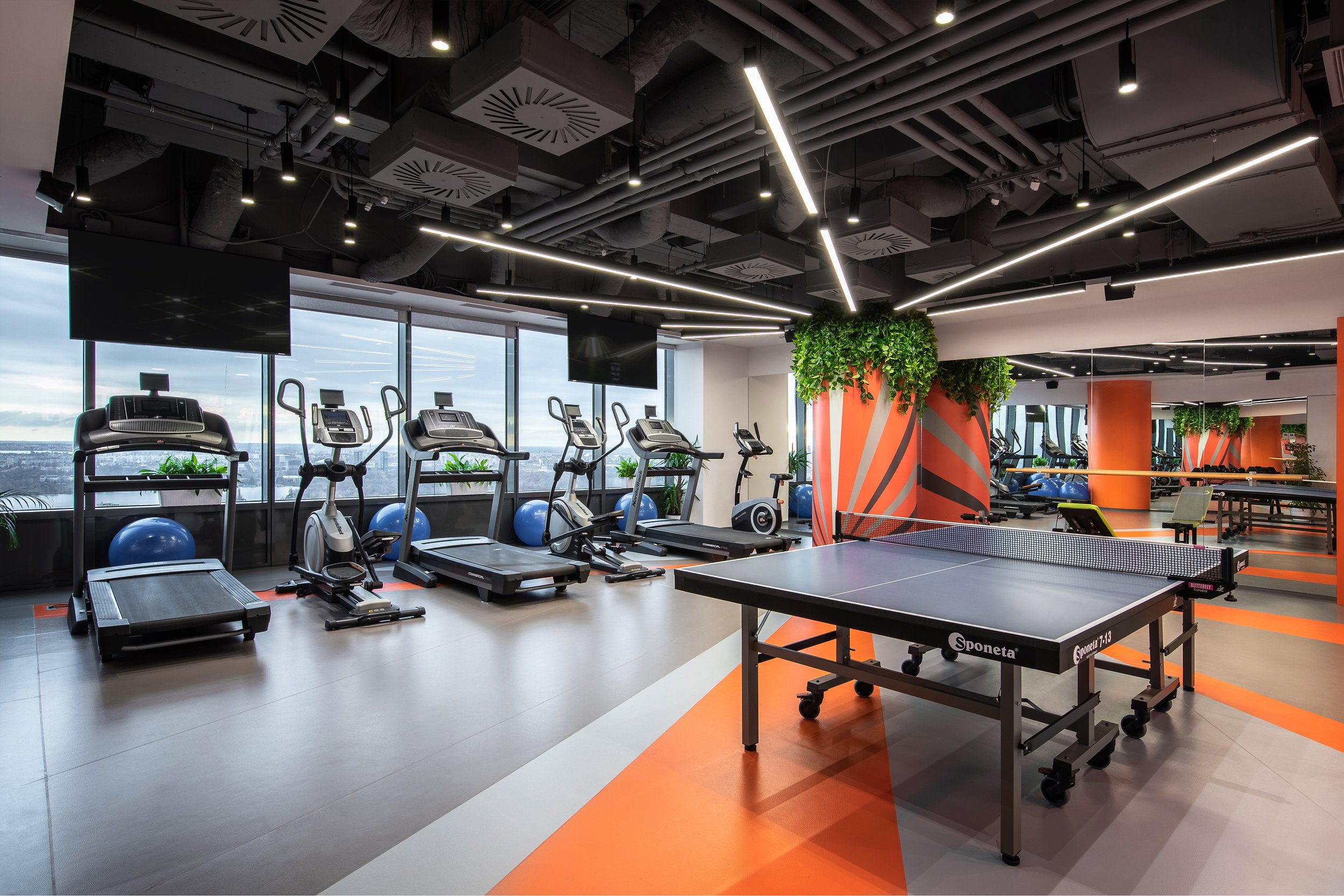 Modern gym with treadmills, exercise bikes, and a ping pong table, featuring large windows, indoor plants, and black ceiling with hanging lights.