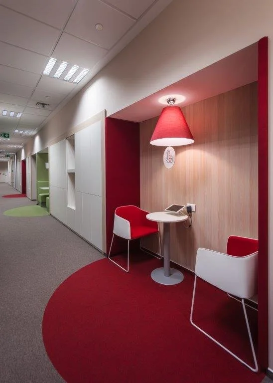 A cozy seating nook with two modern chairs, one red and one white, around a small round table with a telephone. A red lampshade hangs overhead, attached to a wall with wood paneling. The carpet has a red semicircular design matching the wall nook. The hallway stretches into the distance with various colored doorways and decor.