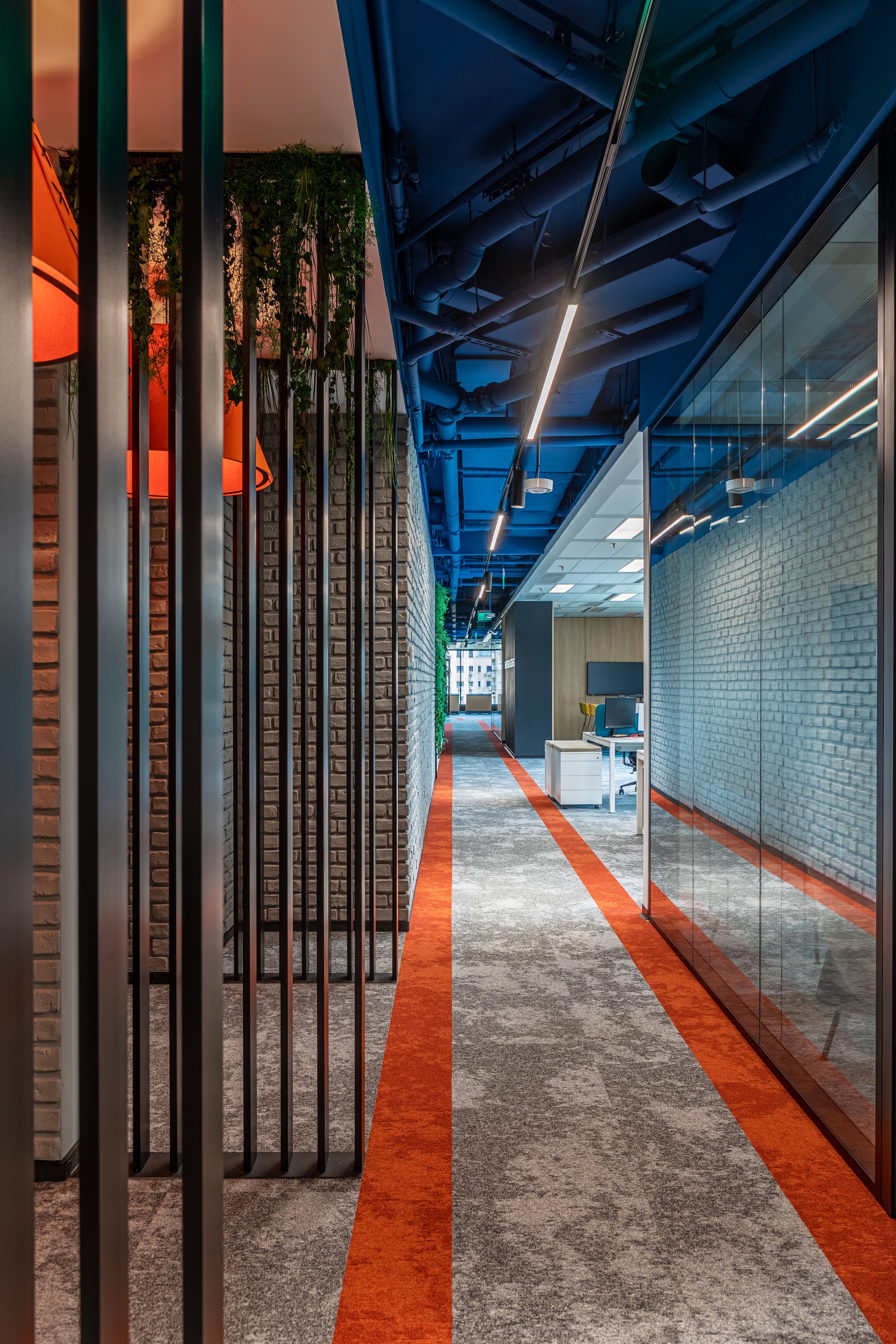 Modern office corridor with colorful carpeting, exposed ceiling pipes, glass walls, and office furniture.